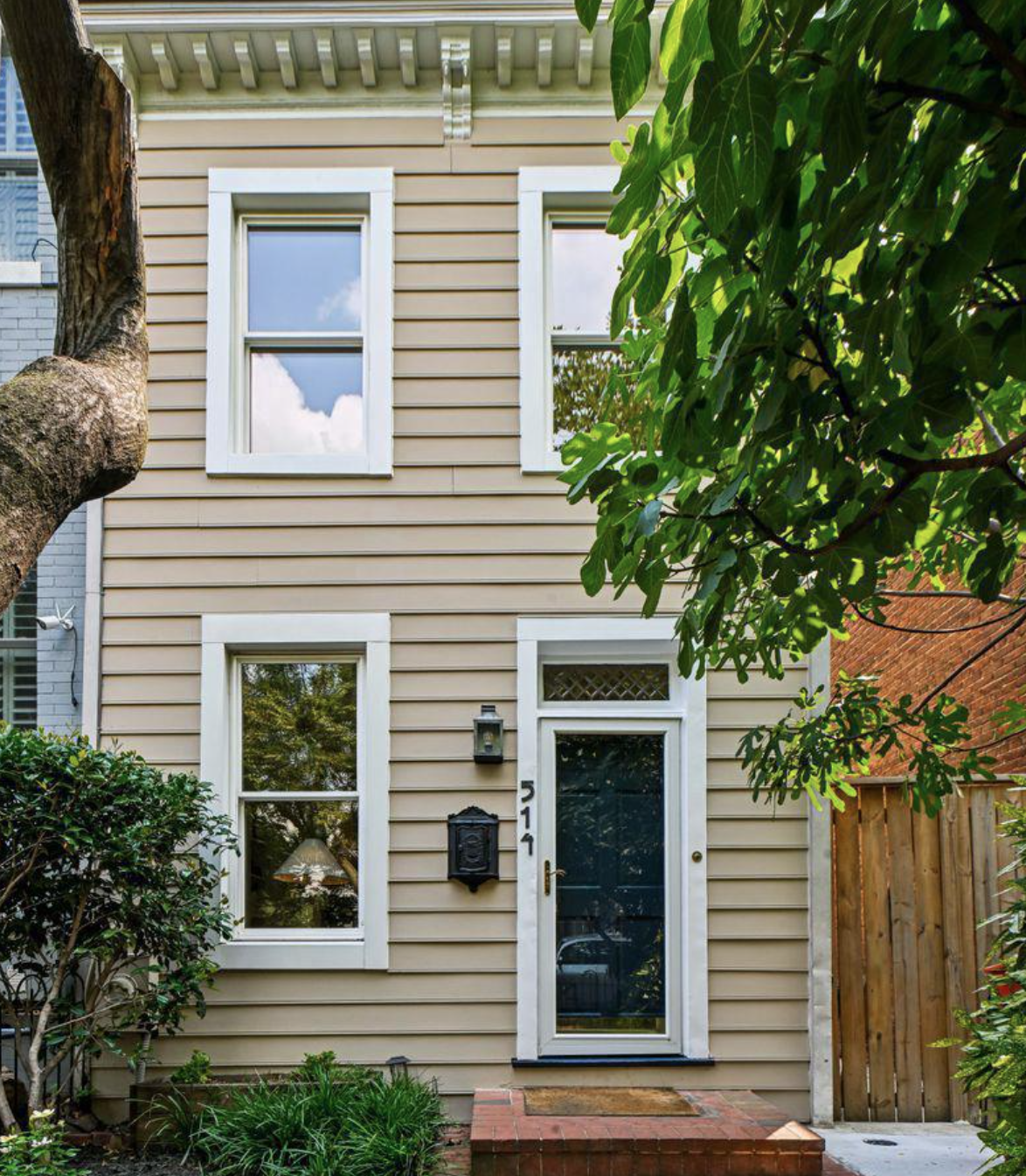 Front view of a beige house with white trim, two upper windows reflecting the sky, and a glass door with house number 514. There is a mailbox next to the door, a small porch rug, and surrounding greenery including a large tree and bushes.