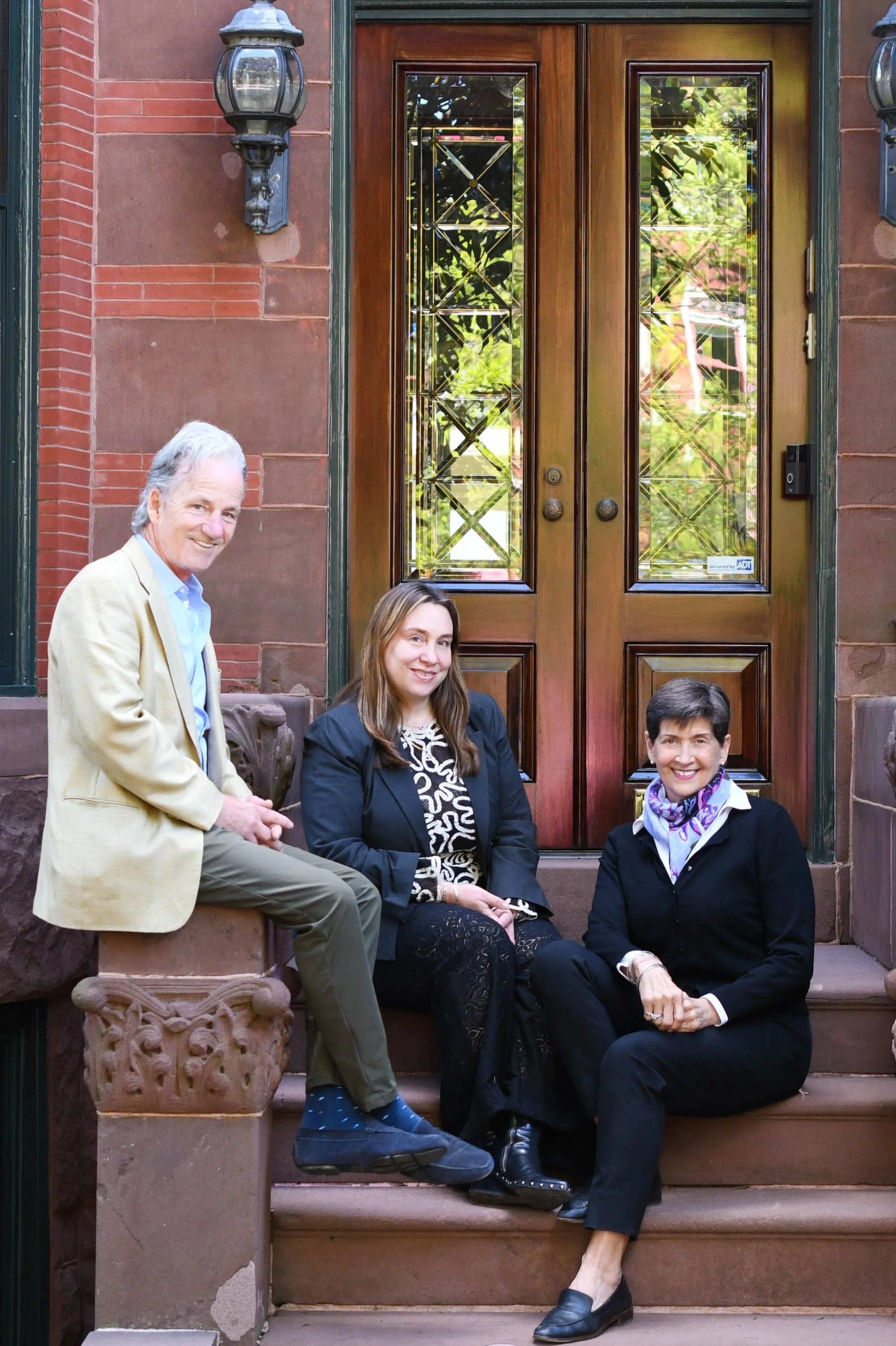 Four people sitting on stone steps outside a building with a wooden door and decorative glass panels, two women sitting, one man standing, and one woman kneeling, all smiling.