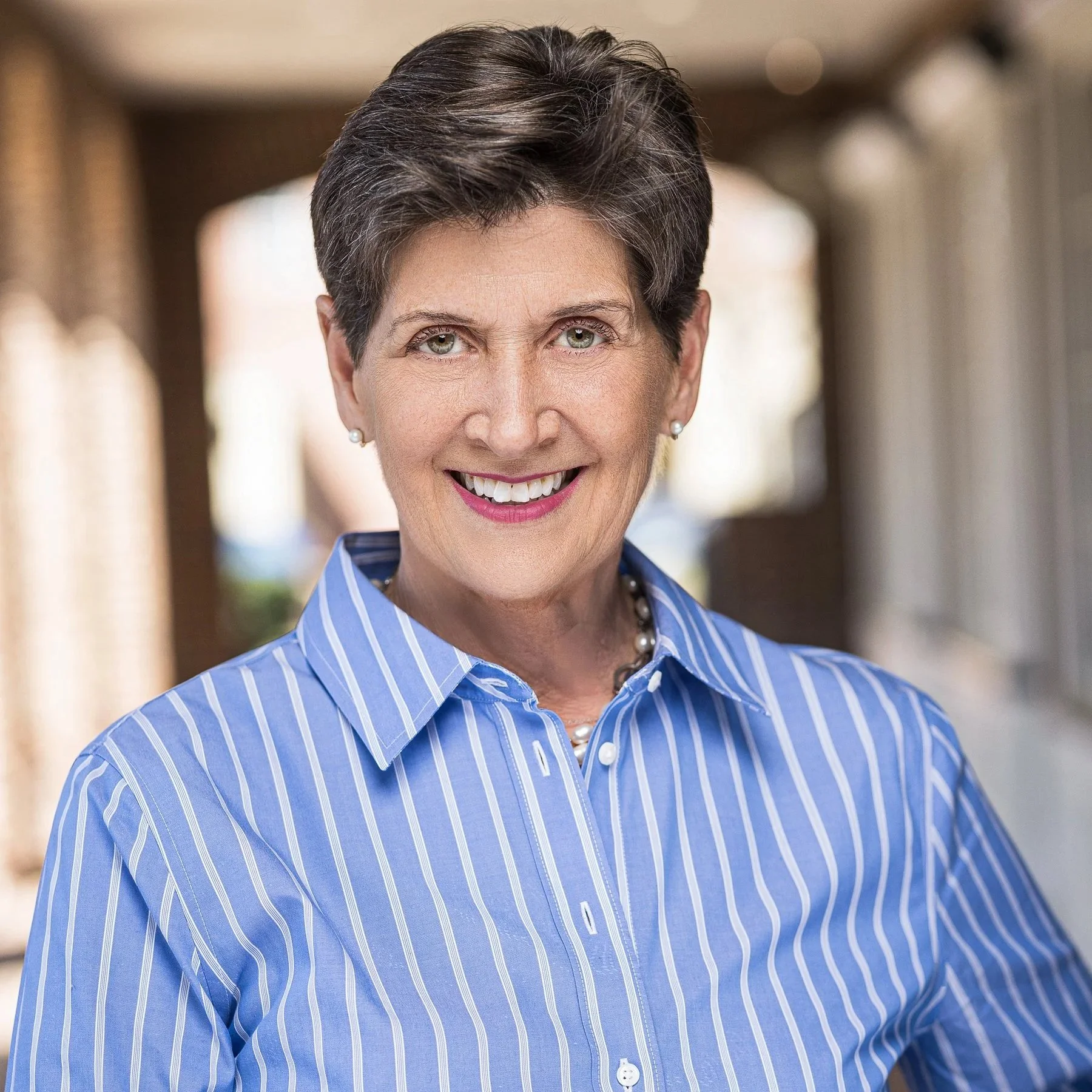Smiling woman with short brown hair and light green eyes wearing a blue and white striped shirt, earrings, and a pearl necklace, standing indoors with blurred background.