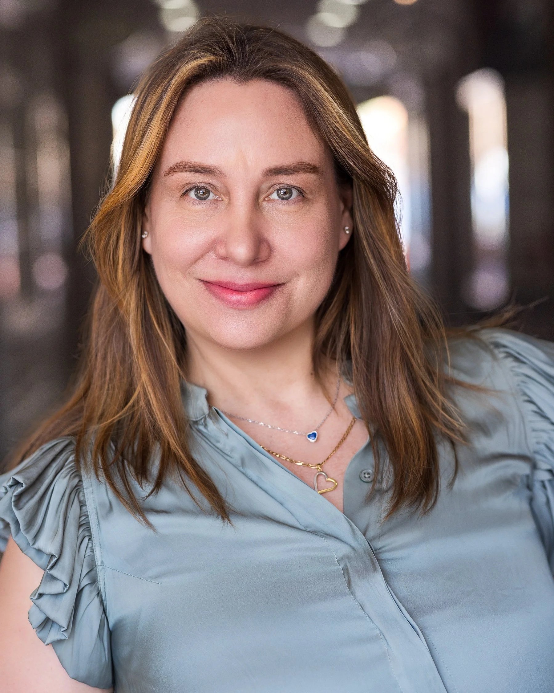 A woman with light brown hair and blue eyes smiling, wearing blue top with ruffled sleeves, layered gold necklaces with heart pendants, and small earrings, standing in a dimly lit space with soft light coming through windows in the background.