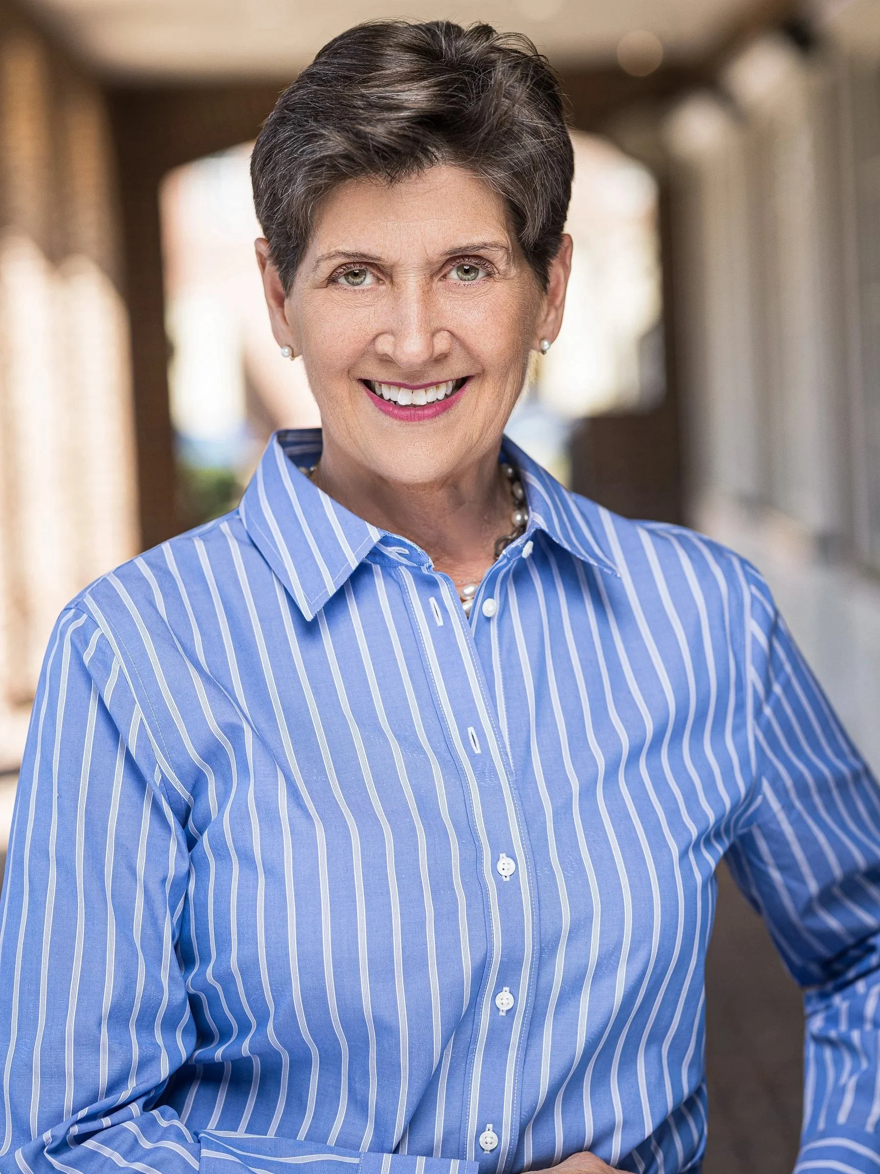 Close-up of a smiling woman with short dark hair, wearing a blue striped button-down shirt and pearl earrings, standing in a brightly lit indoor space.
