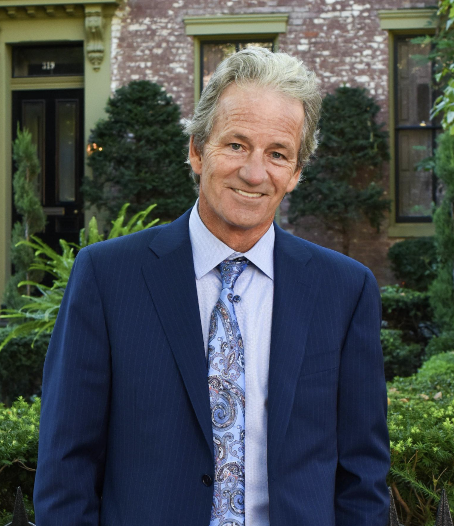 A smiling middle-aged man in a blue suit and patterned tie standing outdoors in front of a brick house with greenery.