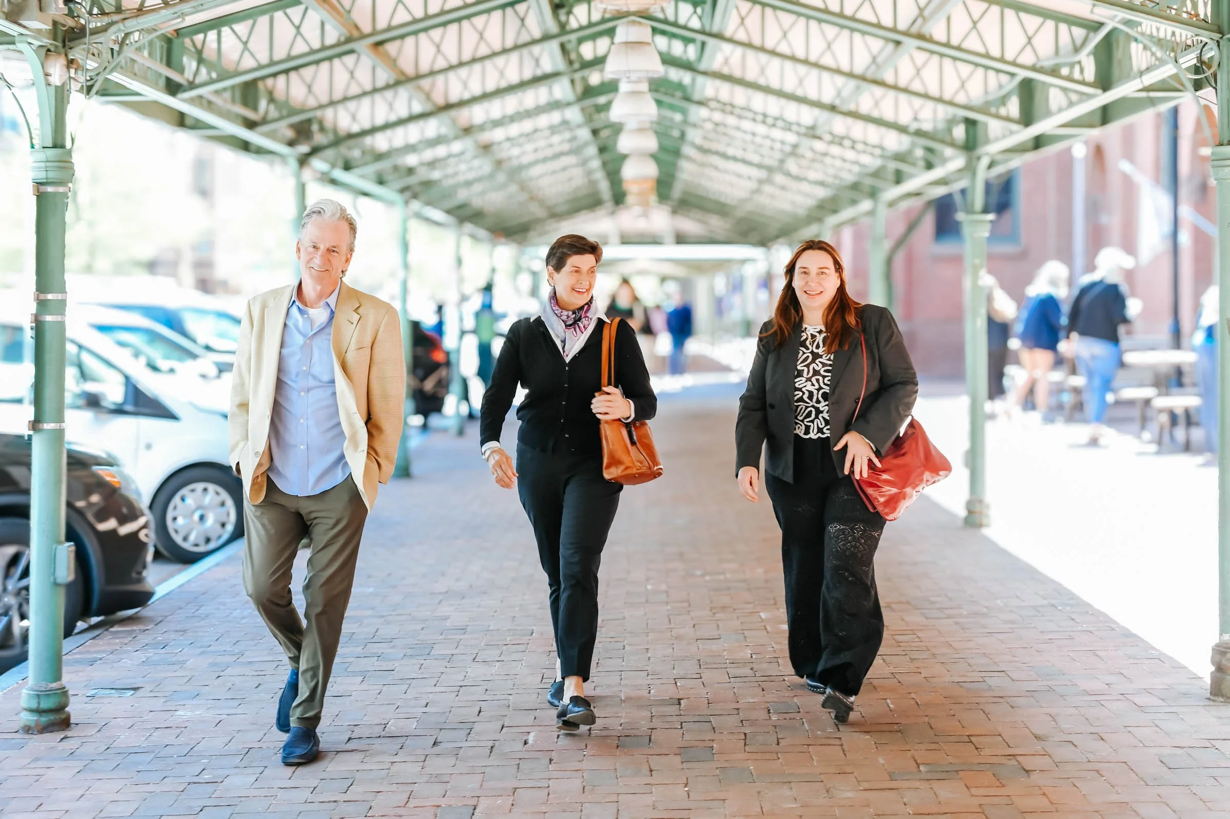 Three pedestrians walking along a covered sidewalk in an urban area. The man on the left is wearing a beige blazer over a light blue shirt. The woman in the middle has short dark hair, a black blazer, and is carrying a brown handbag. The woman on the right has long brown hair, a black blazer, a patterned shirt, and is carrying a red bag. There are parked cars on the left and buildings on the right in the background.