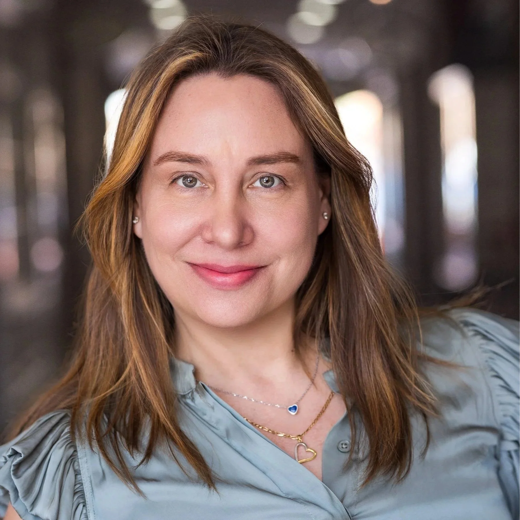 Close-up portrait of a smiling woman with long brown hair, wearing a blue shirt, layered necklaces, and earrings, in a softly blurred background.