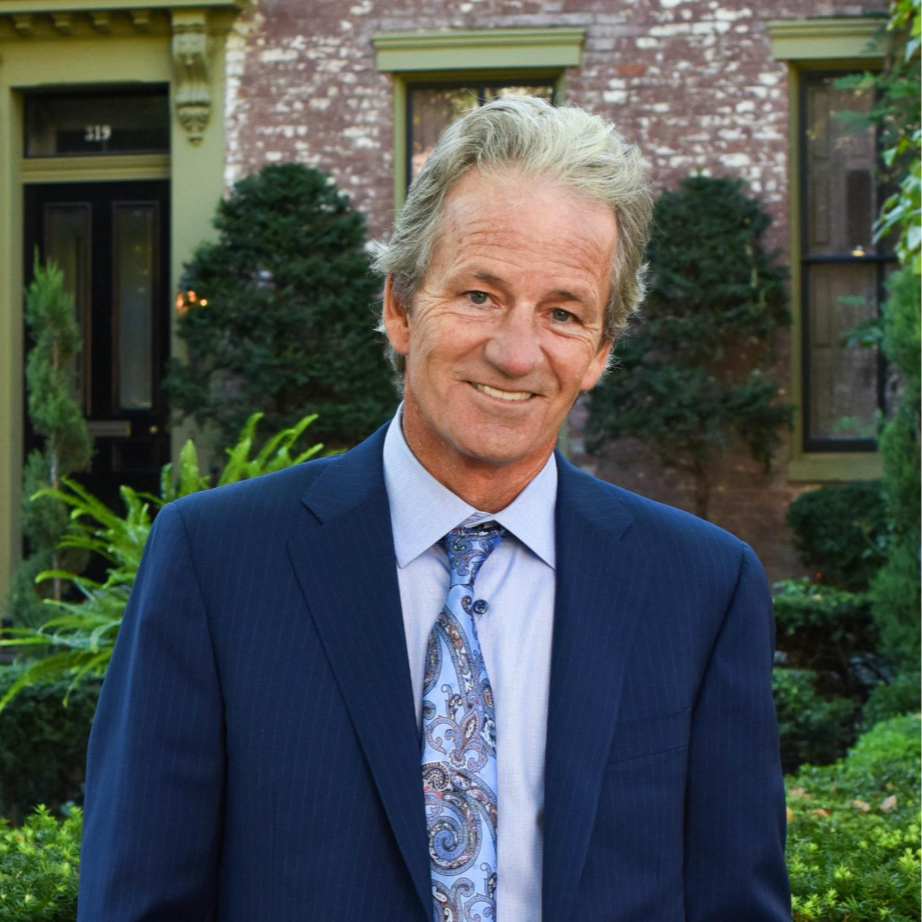 A smiling middle-aged man with gray hair, wearing a blue suit, light blue shirt, and patterned tie, standing outside in front of a brick building with greenery.
