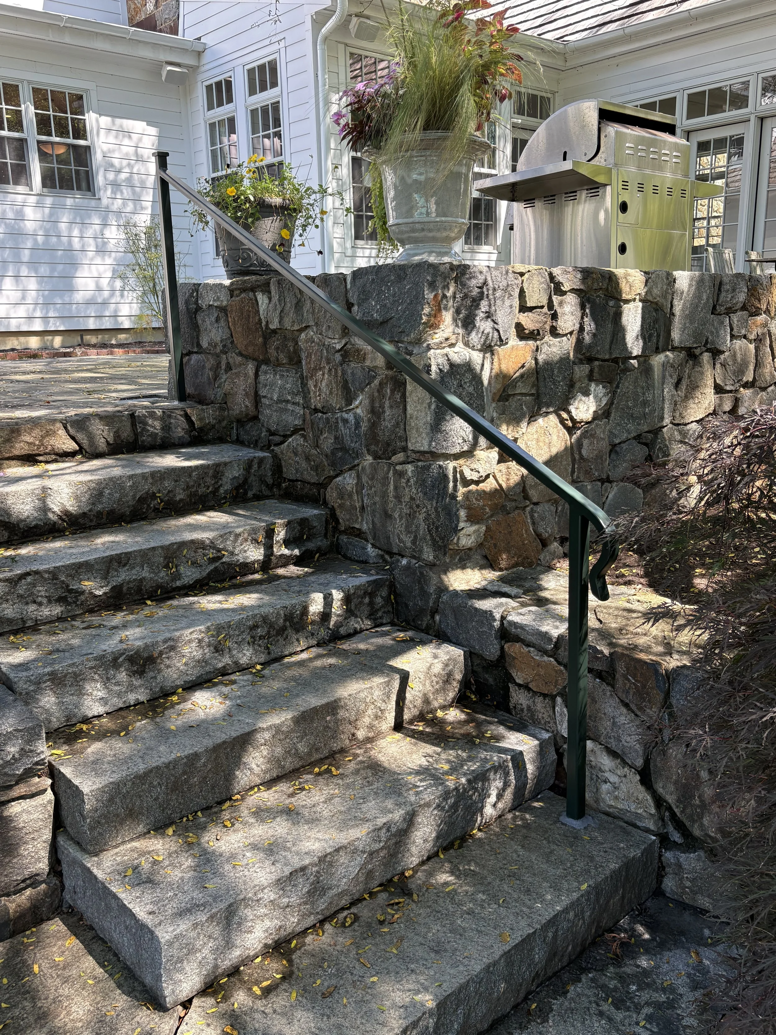 Stone steps with a metal railing leading up to a patio area with plants and a stainless steel barbecue grill next to a white house with multiple windows.