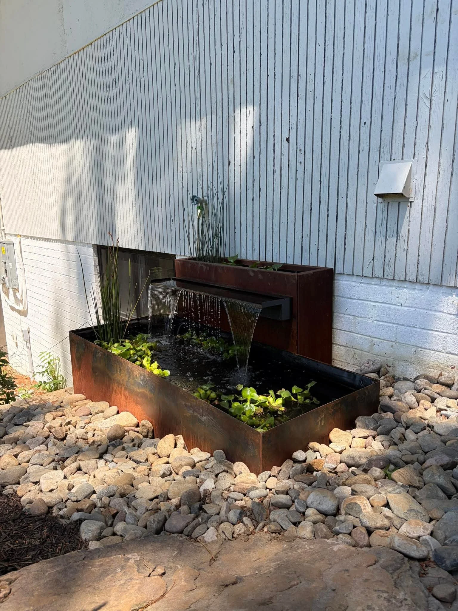 A rectangular Corten steel water feature with floating lily pads and small plants, surrounded by rocks, with a white wooden exterior wall and a window with plant below it in Baltimore MD