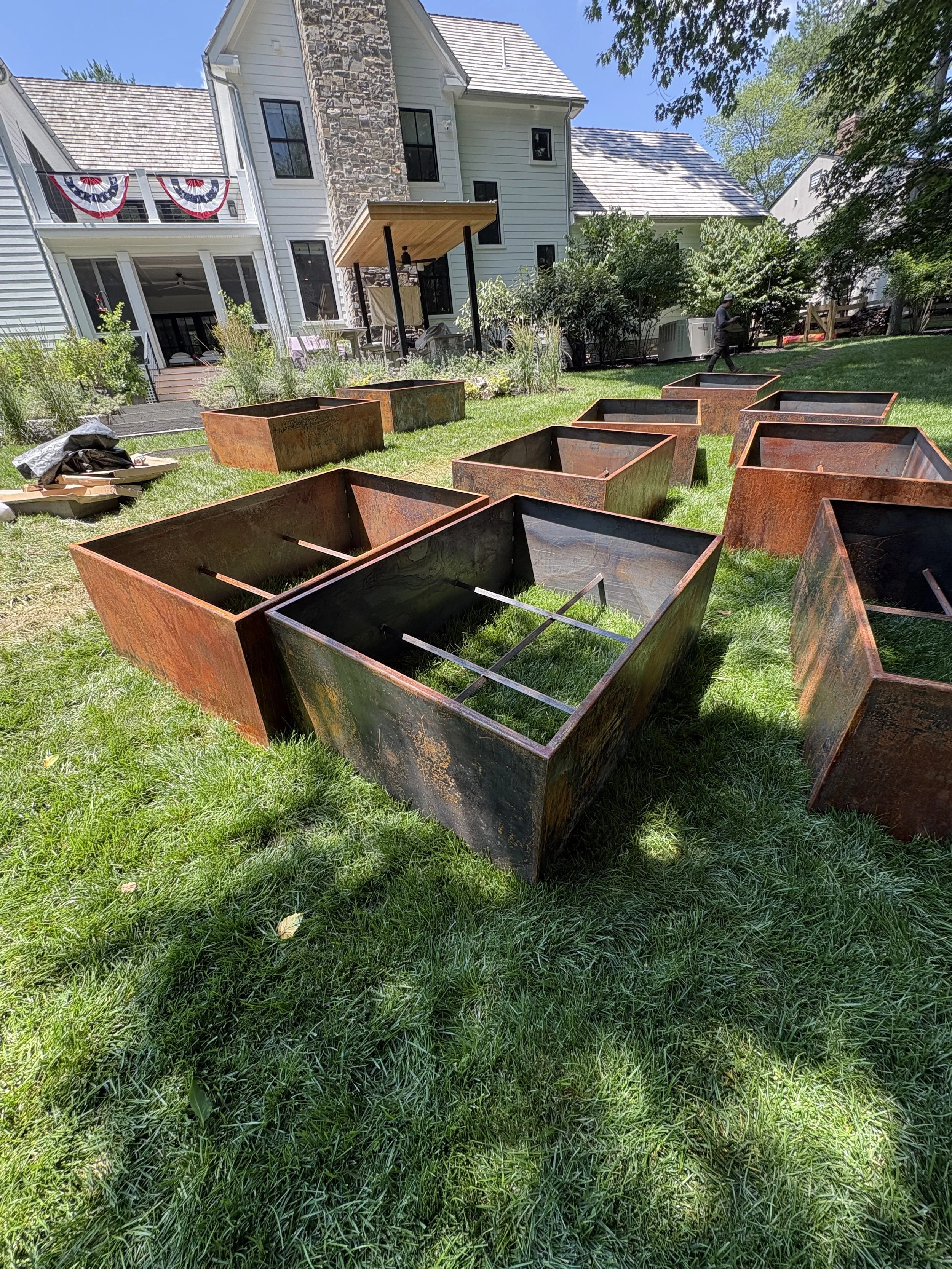 A backyard scene with Corten steel planters on green grass, a house with white siding and a stone chimney, and a person walking near trees in Chestnut Hill Philadelphia