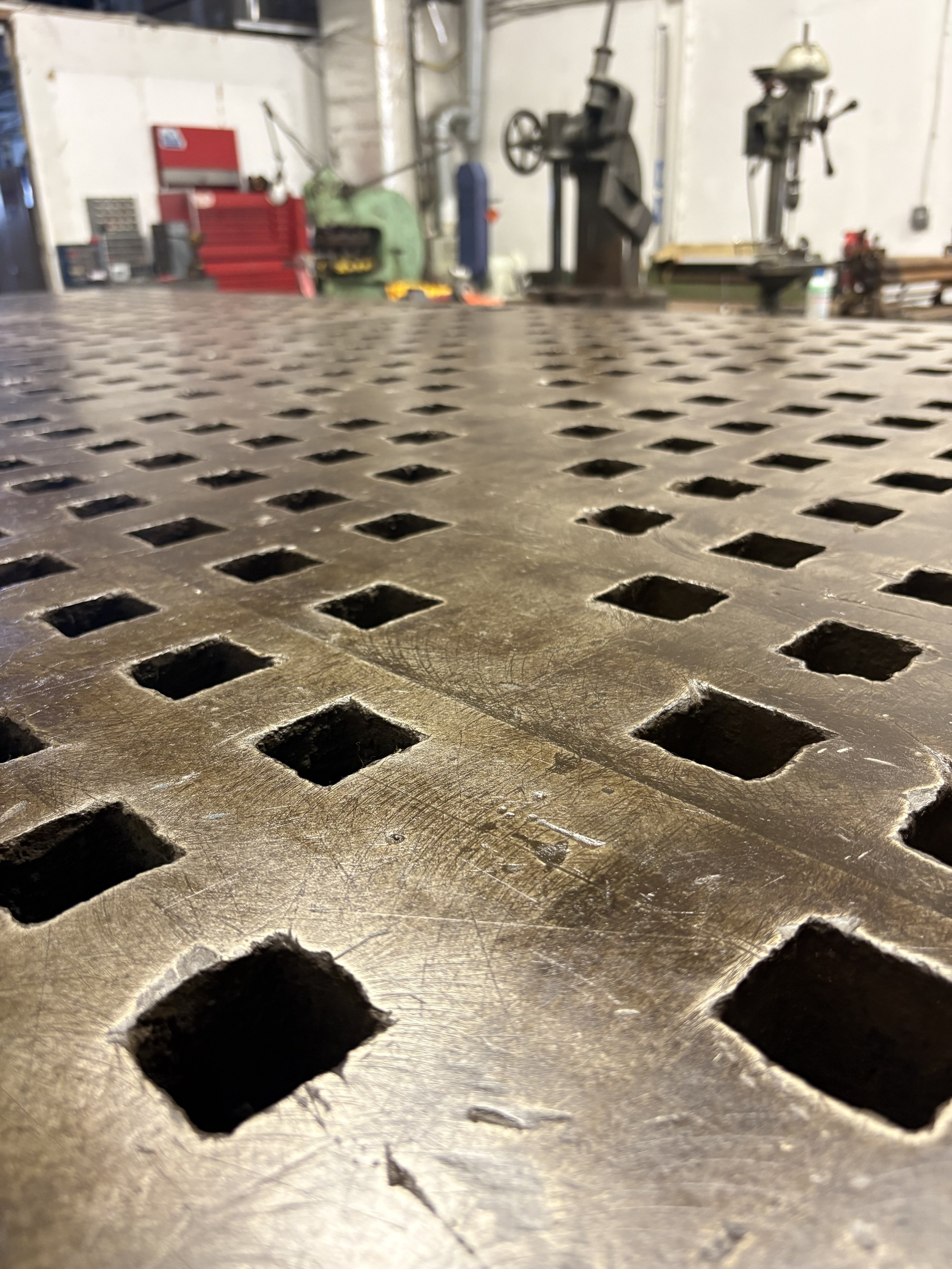 Close-up view of a vintage welding fixture table with a perforated surface, typically used for industrial or workshop purposes, with blurred machinery and tools in the background.