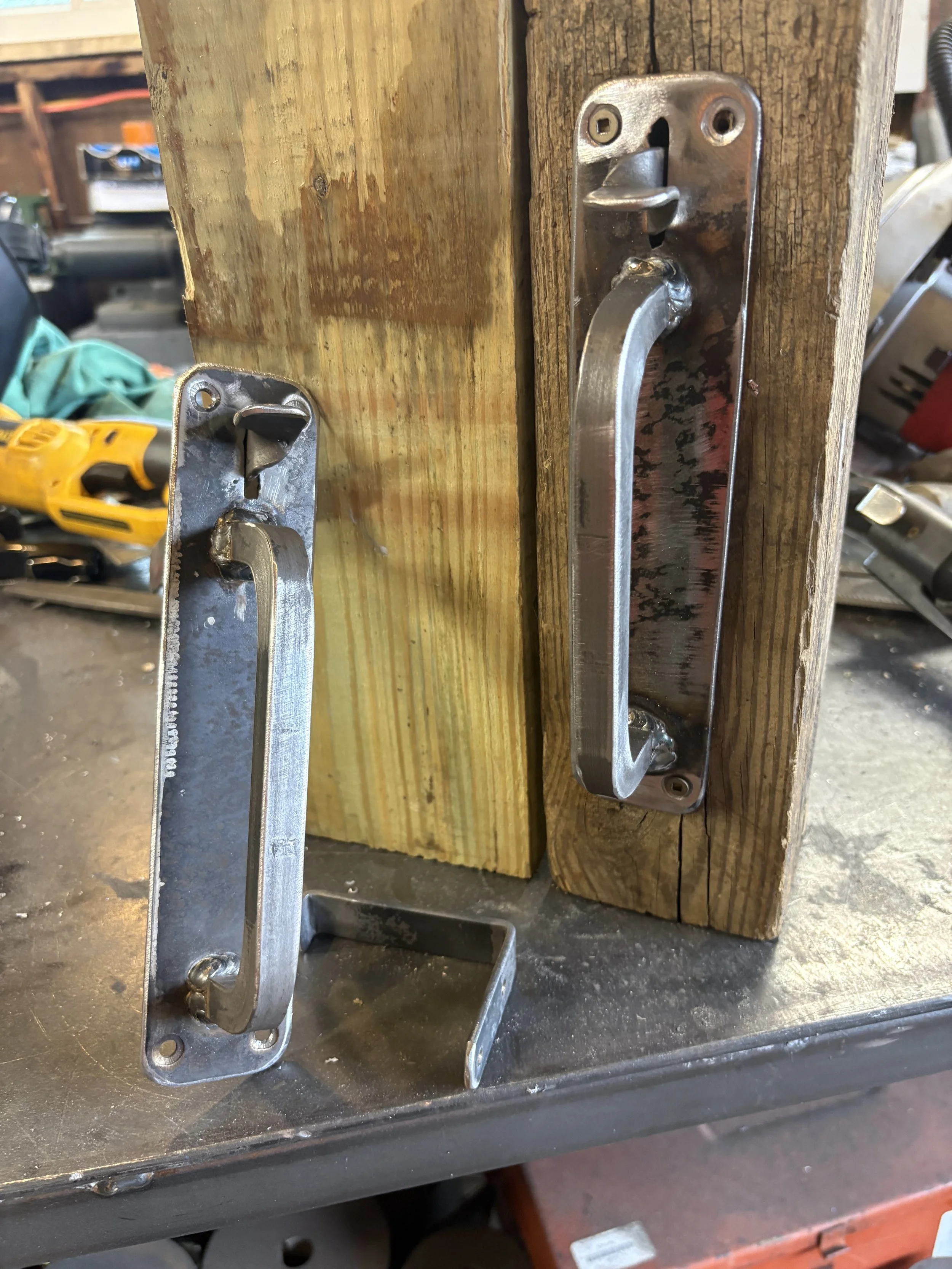 Metal gate handles attached to a wooden plank in a workshop setting.