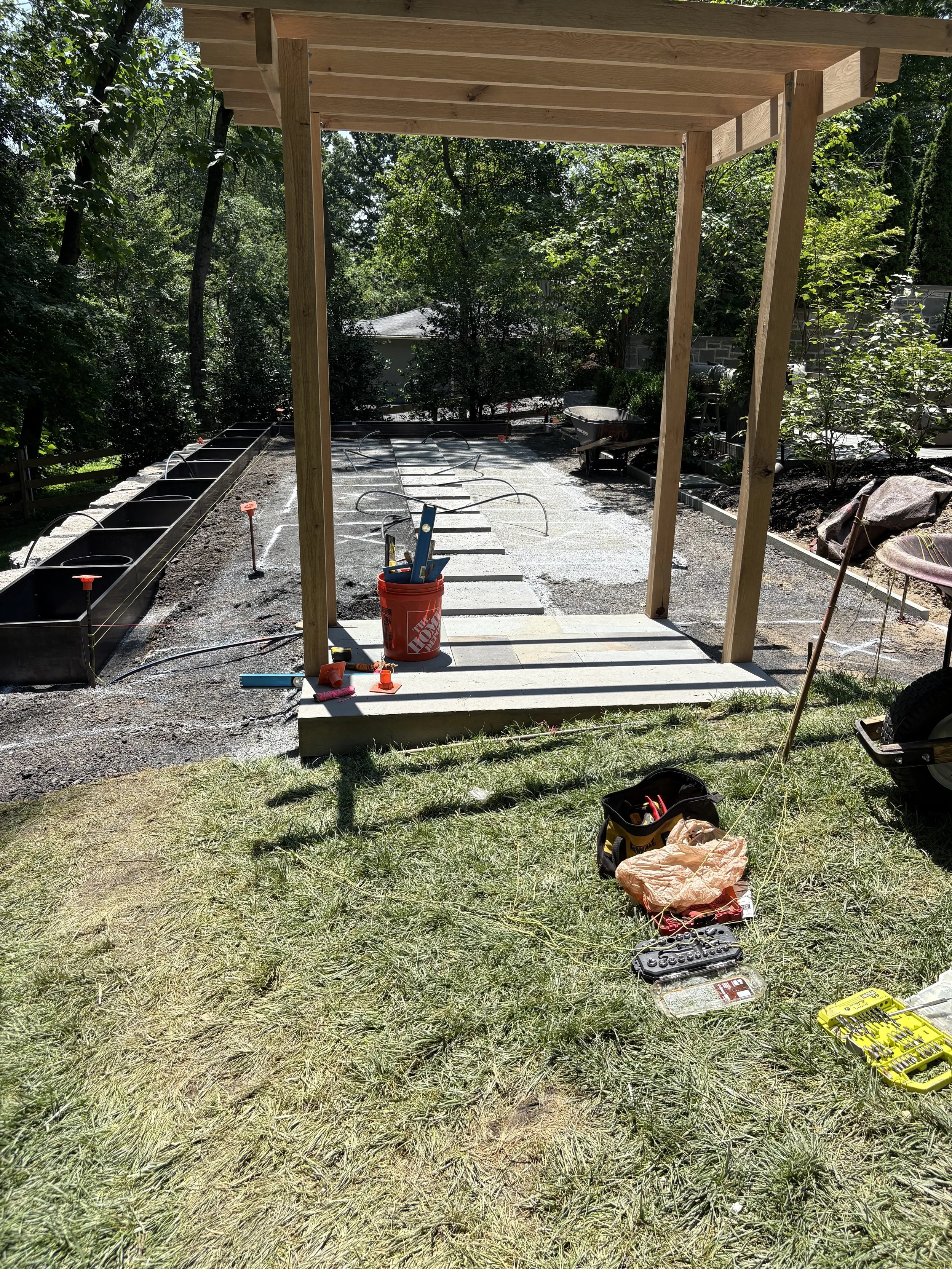 Construction site for a custom garden with wooden frame and tools, including a toolbox, a wheelbarrow, and construction materials, on a grassy area with trees in the background. 