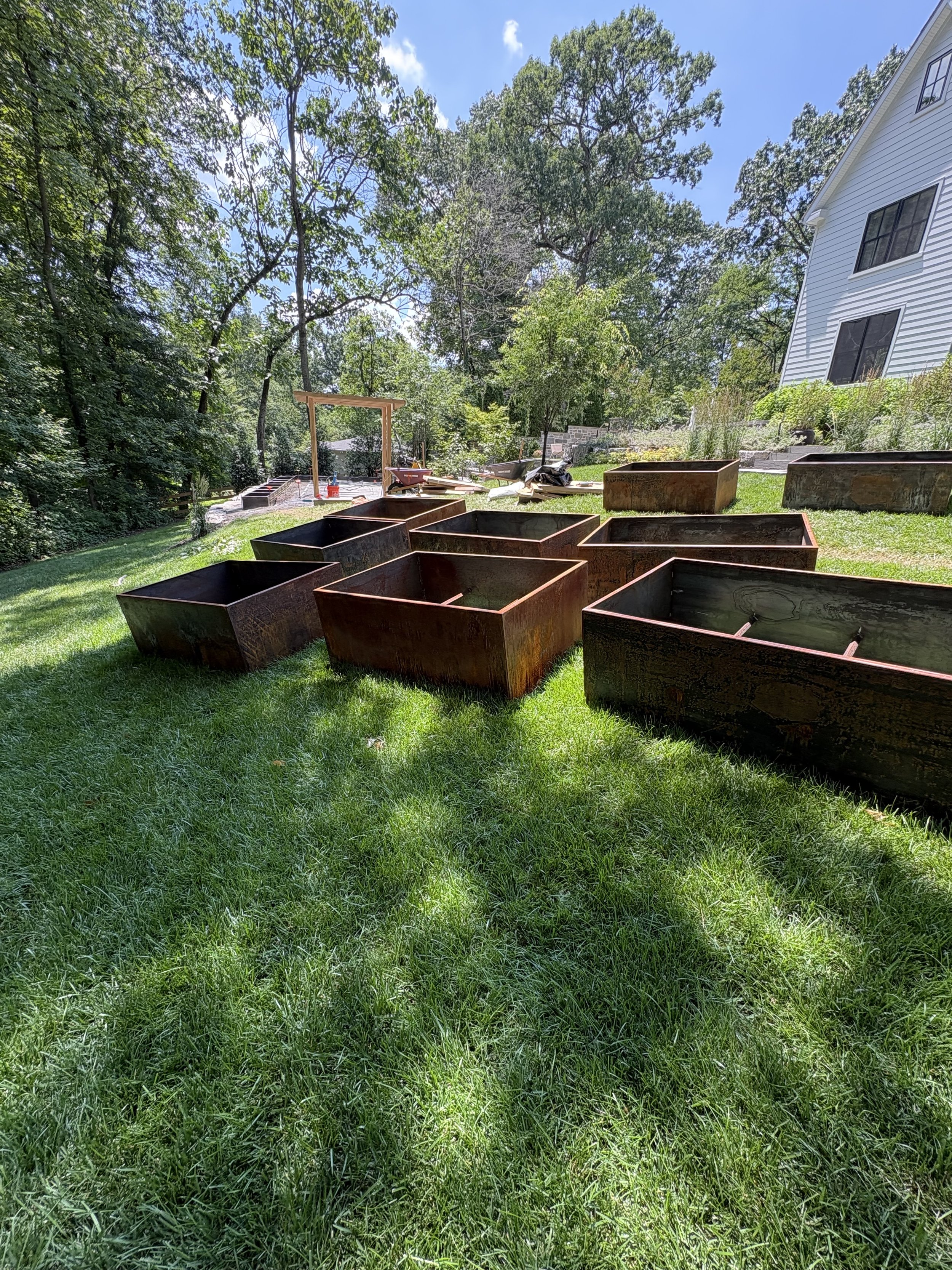 A backyard with a green lawn, trees, and several Corten metal planters showing patina. There are construction tools and materials, including wheelbarrows and a wooden frame, indicating ongoing landscaping work. Chestnut Hill PA