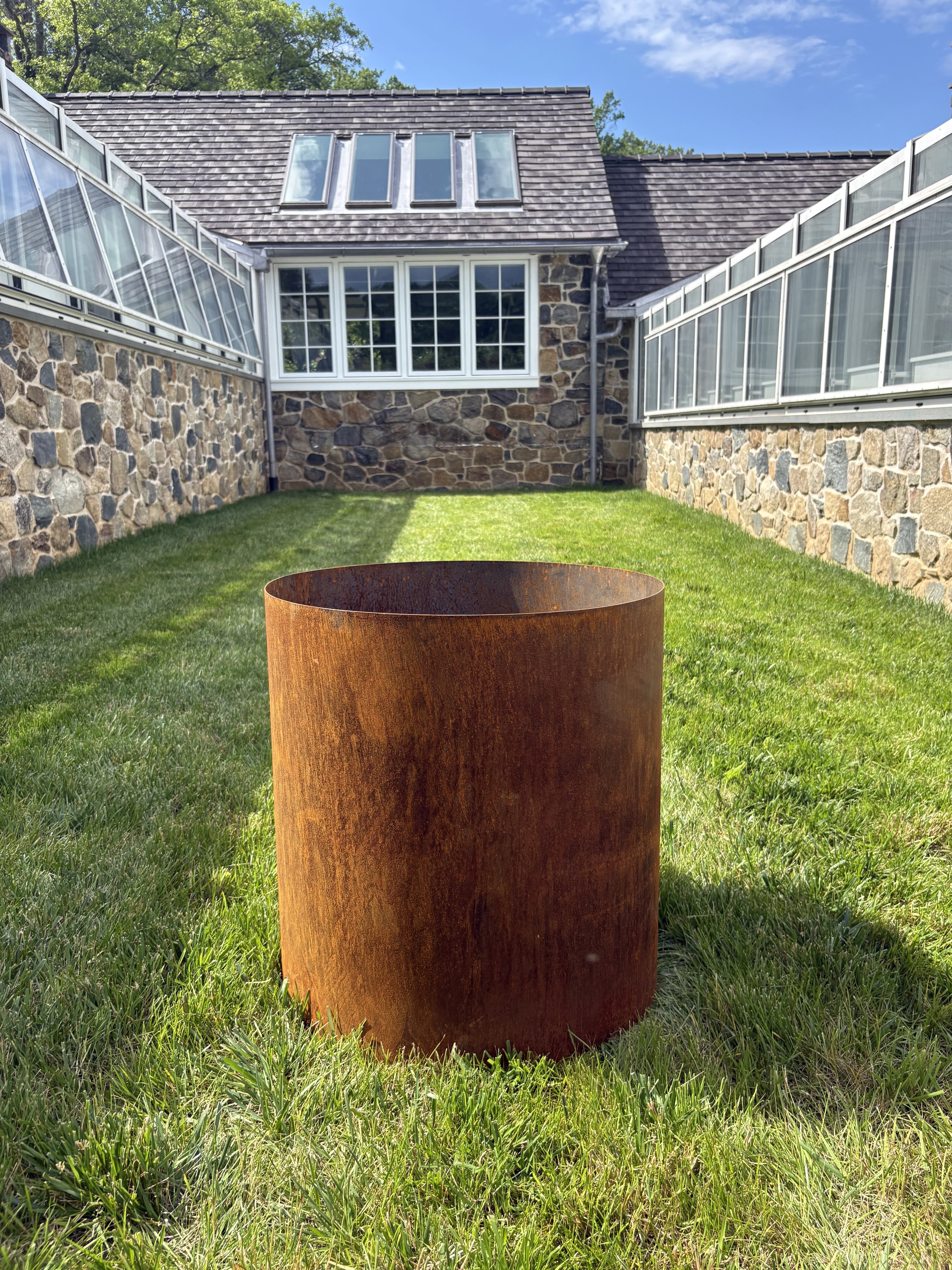 A corten steel planter placed on lush green grass behind a greenhouse with white-framed windows, a sloped roof with skylights, and glass-enclosed rooms on either side. Clear blue sky above.