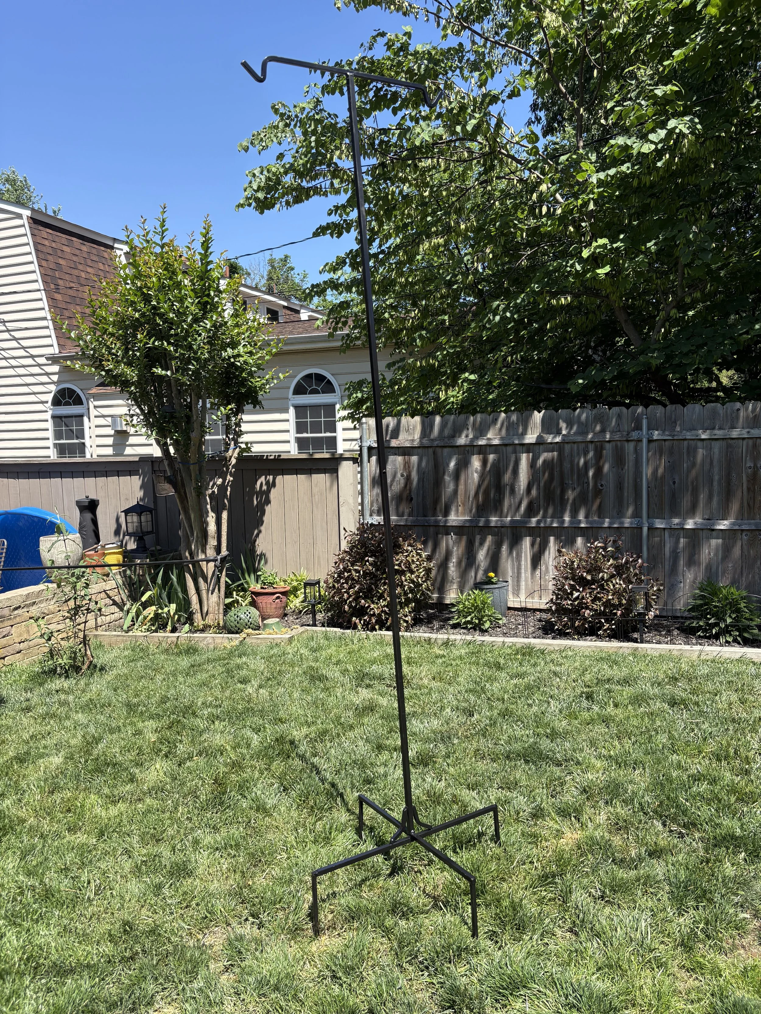 A backyard with a grassy lawn, small garden, and trees. A tall, curved black metal shepherds hook bird feeder is in the center. In the background, there is a house with cream-colored siding, arched windows, and a fenced backyard.