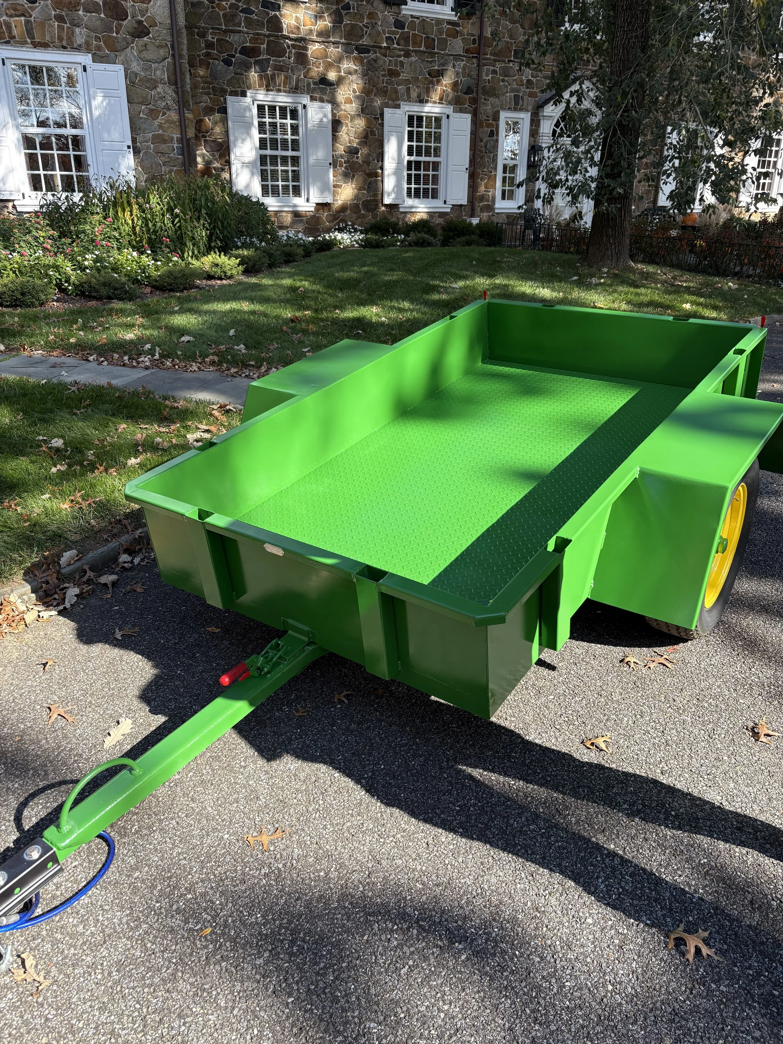 A bright green utility trailer parked on a paved driveway in front of a house with a stone facade, white window shutters, and a large tree providing shade.