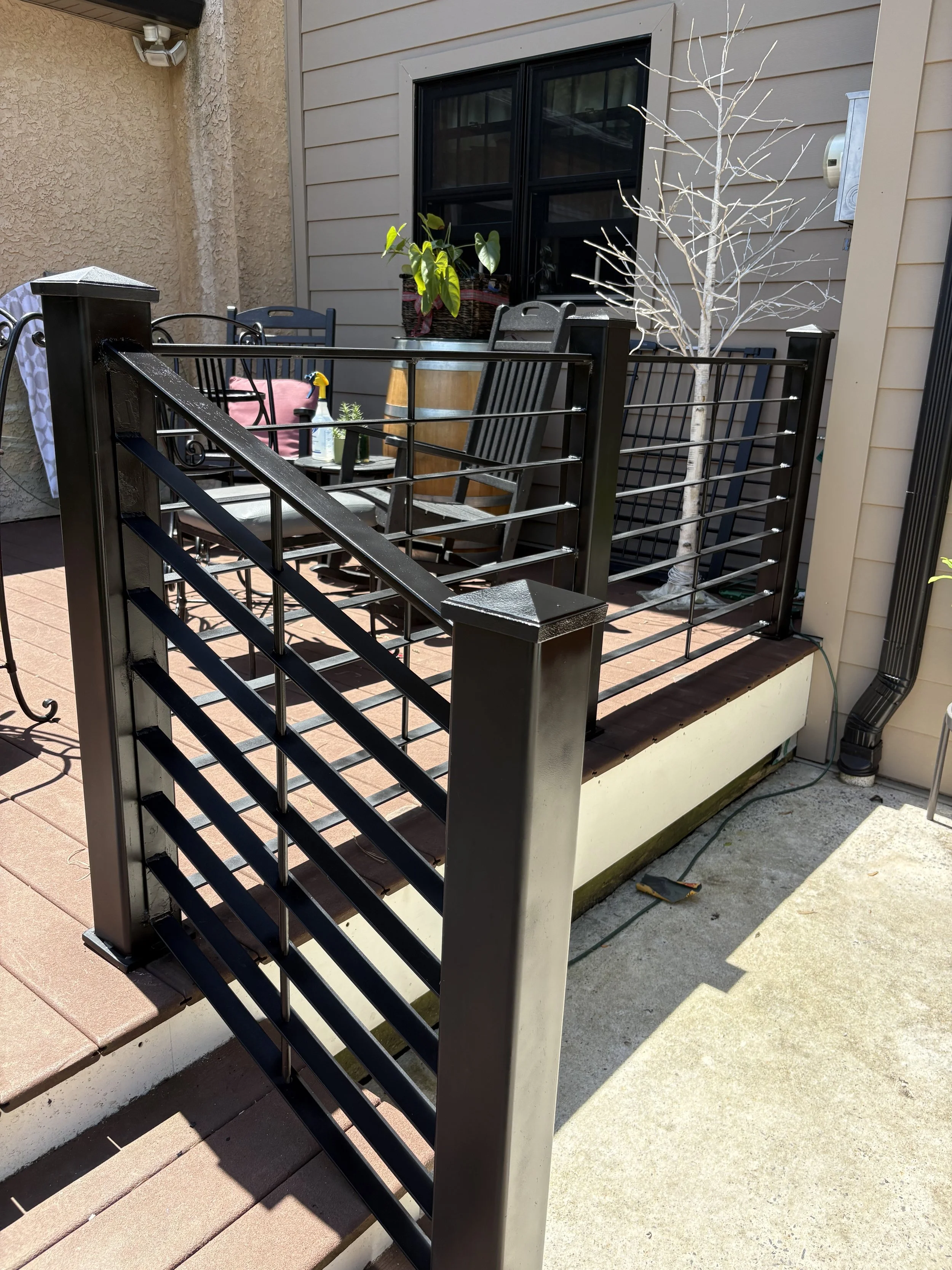 View of a small outdoor patio with a black metal railing, two chairs, a small table with plants, and a leafless tree. The patio is next to a beige stucco wall and gray siding, with a window and a gray downspout visible.
