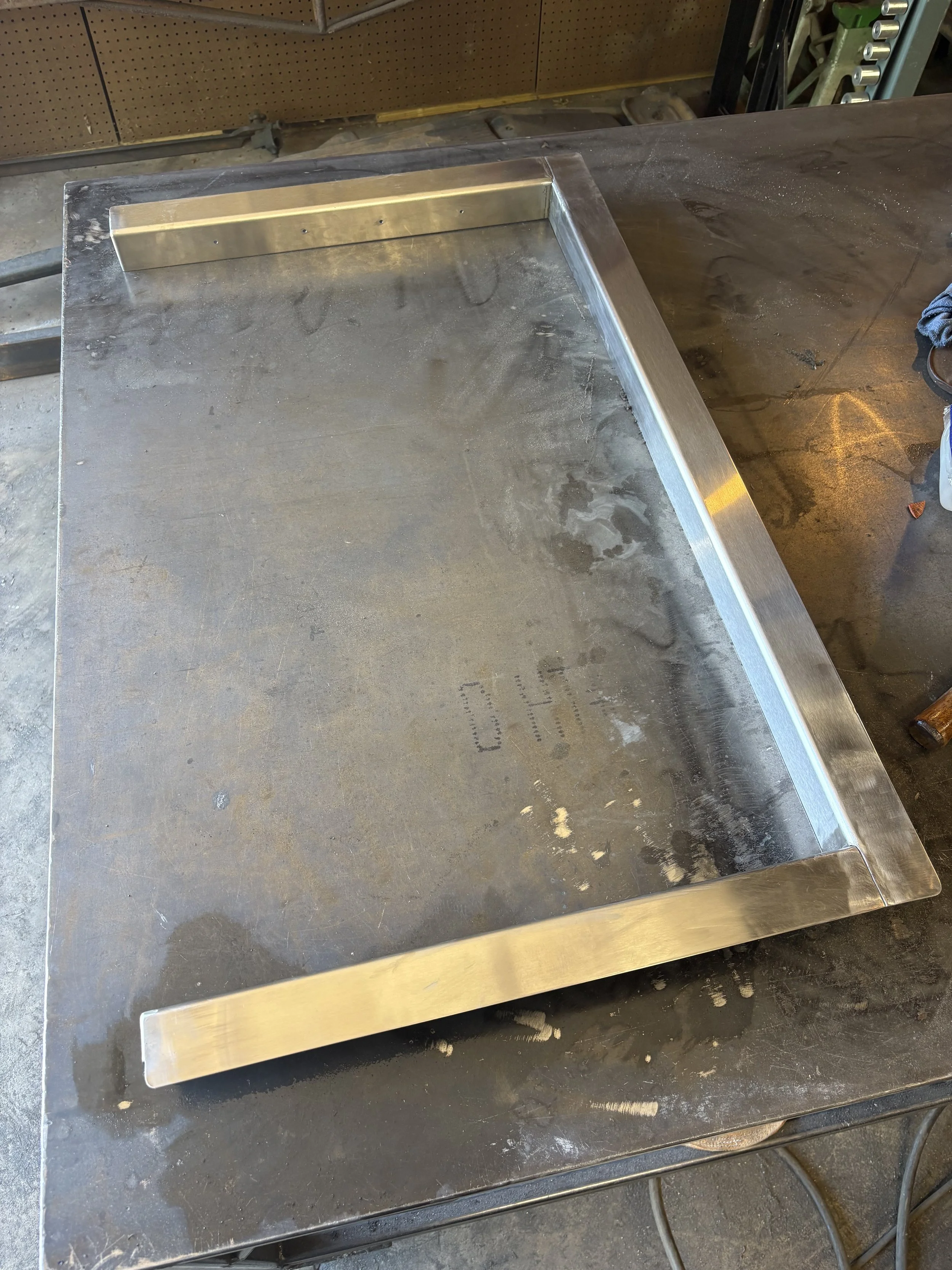 A stainless steel grill surround in a workshop or garage, sitting on a steel work table. Background shows a pegboard with tools and equipment storage, and some tools are visible on the right side.