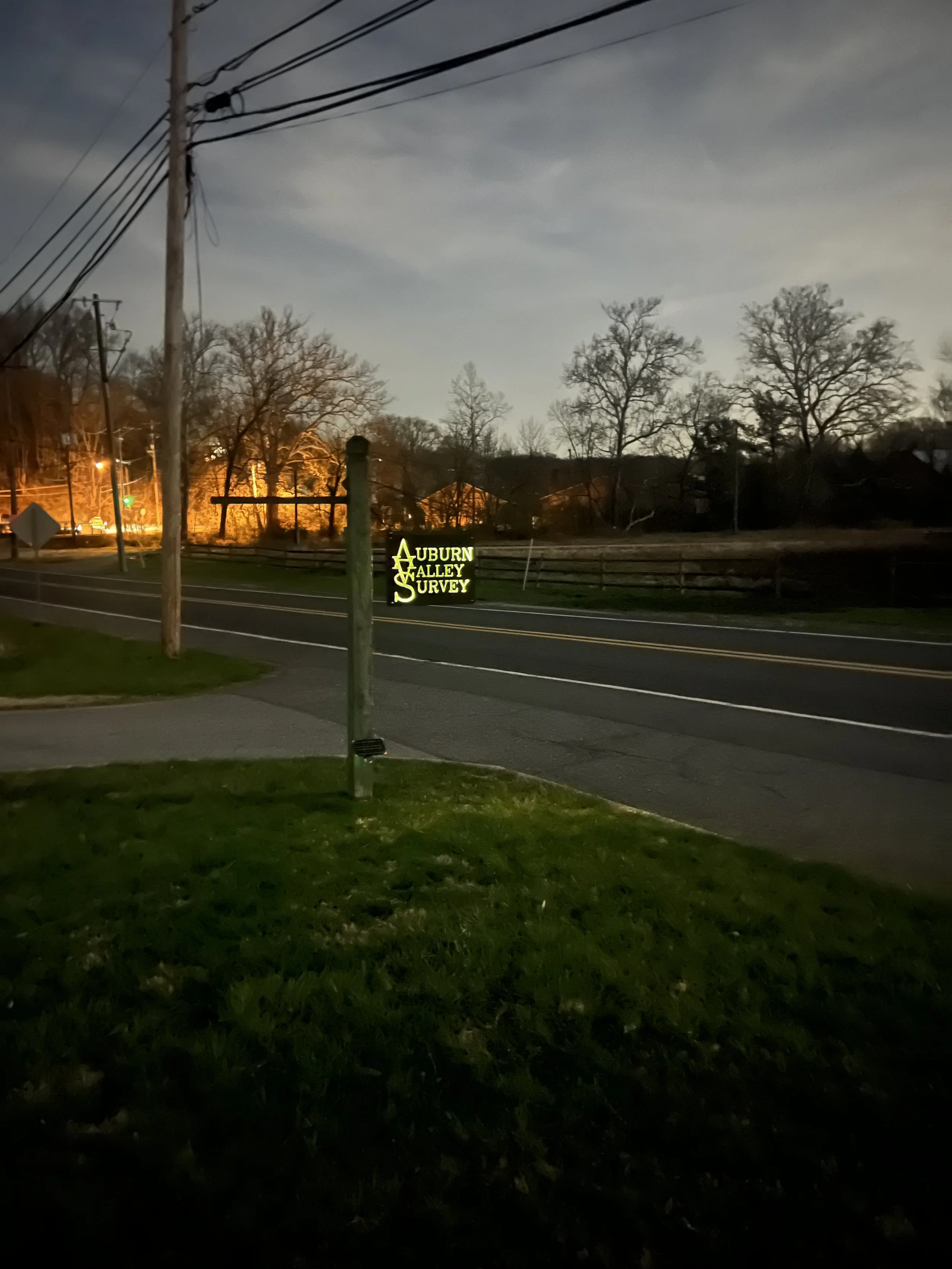 Nighttime scene of a street with utility poles and power lines, trees in the background, and a lit sign that reads 'AUBURN VALLEY SURVEY' near the sidewalk.
