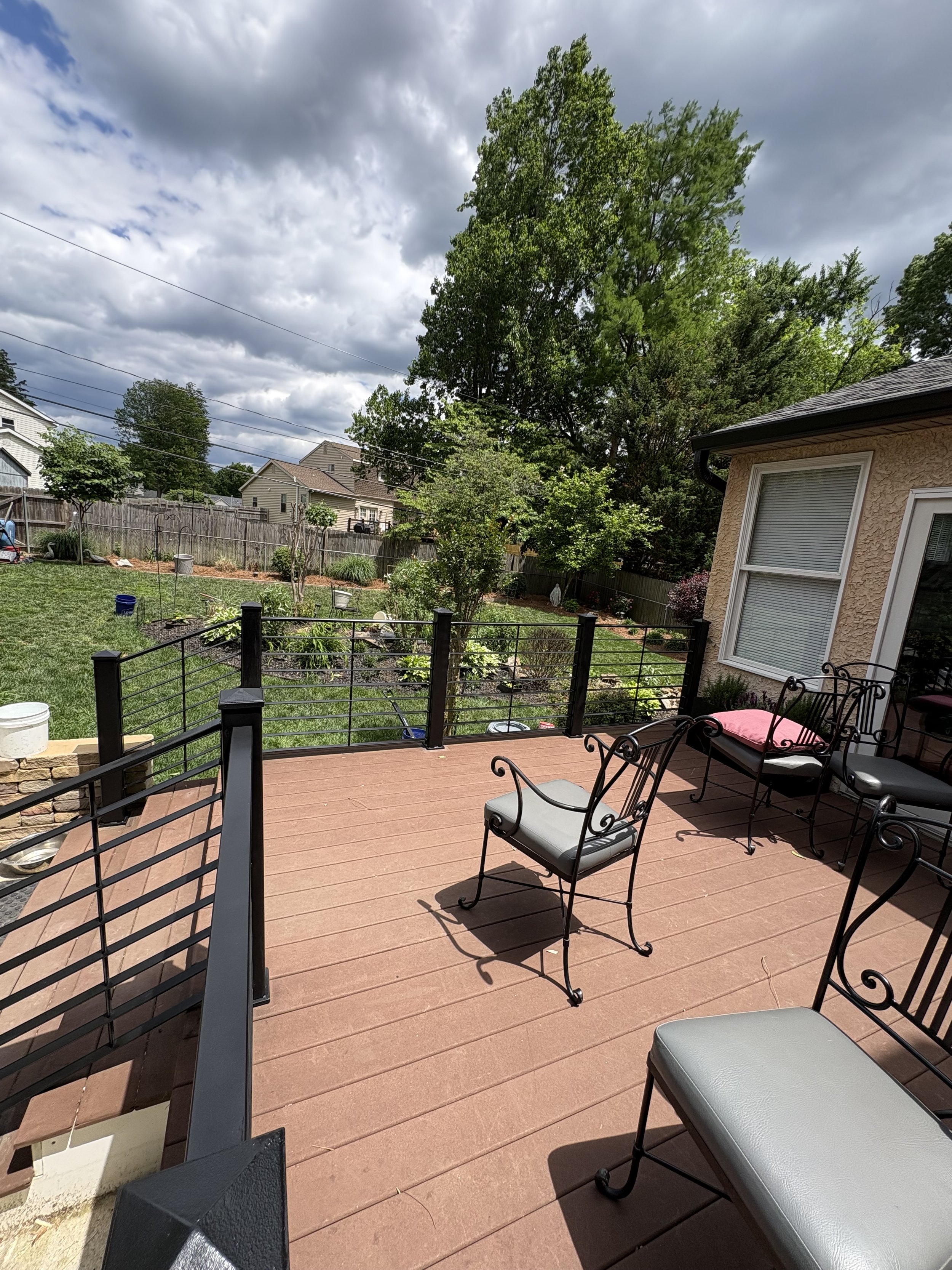 View of a backyard patio area with metal chairs and a table on a wooden deck. The yard has a green lawn, trees, shrubs, and a wooden fence. Overcast sky with clouds. There is a custom metal railing on the patio area