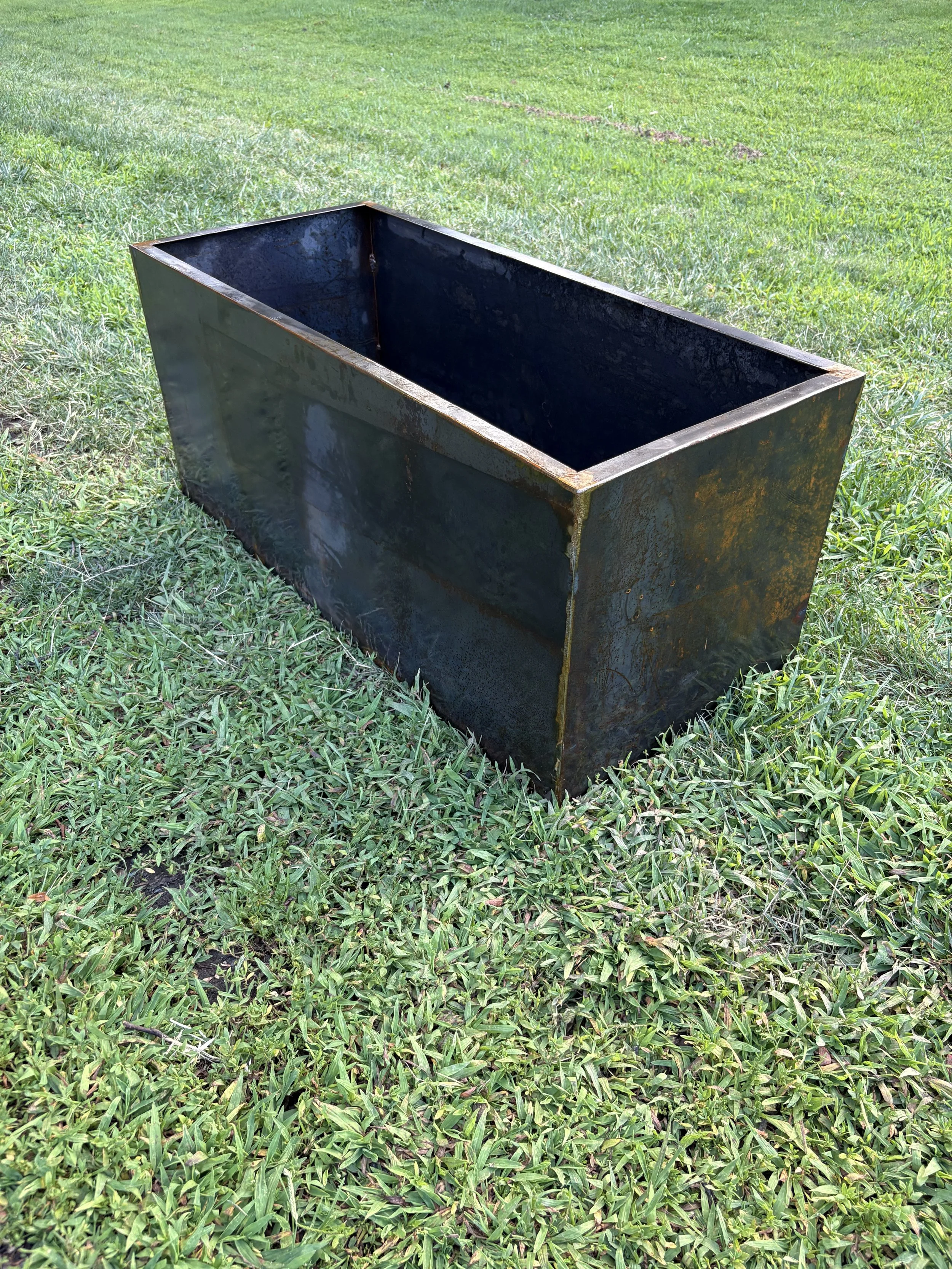 A large rectangular Corten steel metal planter placed on a grassy field.