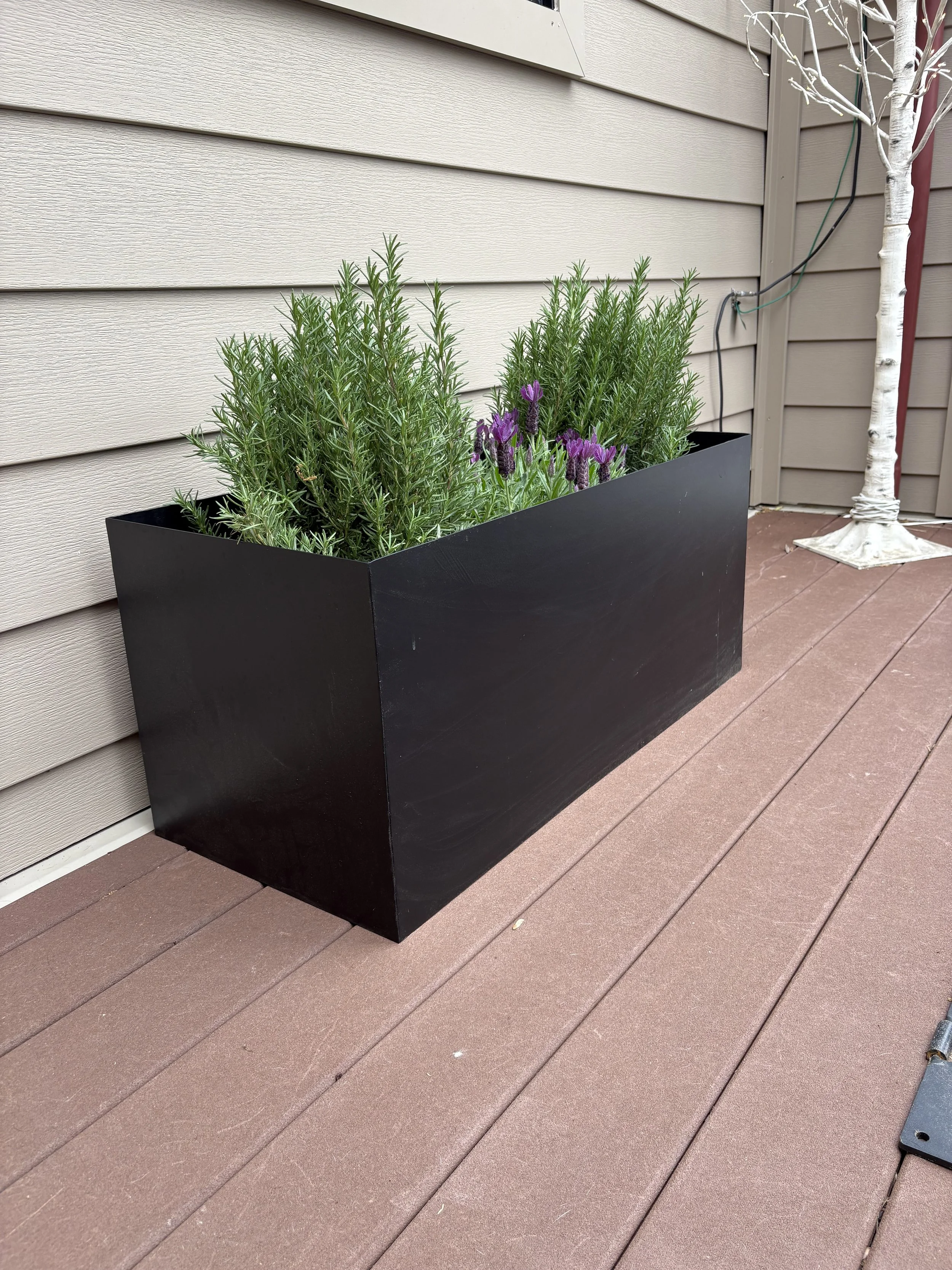 A black rectangular planter with green lavender and purple lavender flowers, placed on a brown wooden deck next to a beige house wall.
