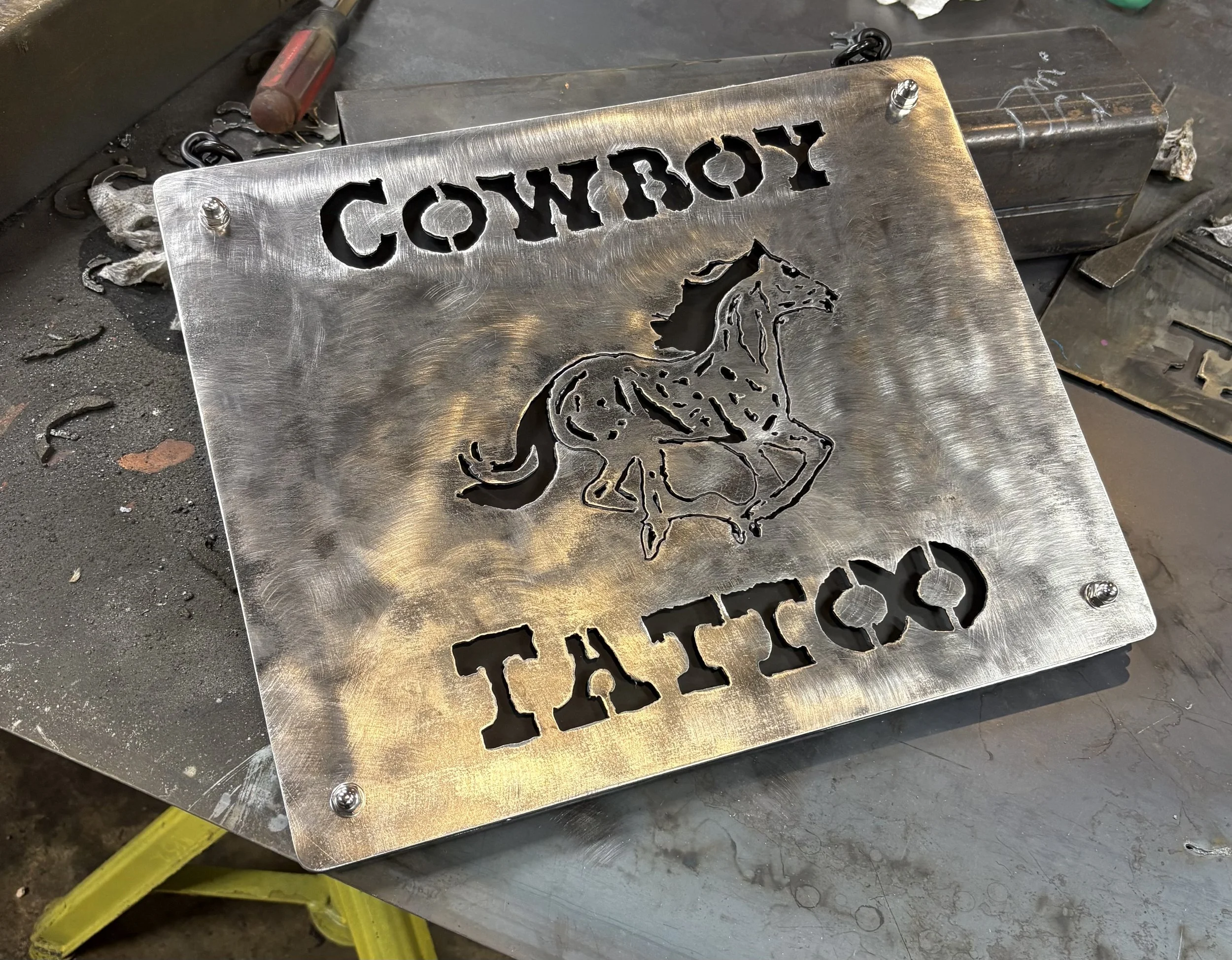 A metal sign with the word 'COWBOY TATTOO' cut out at the top and an illustration of a cowboy riding a horse below it. The sign is on a workbench with tools around