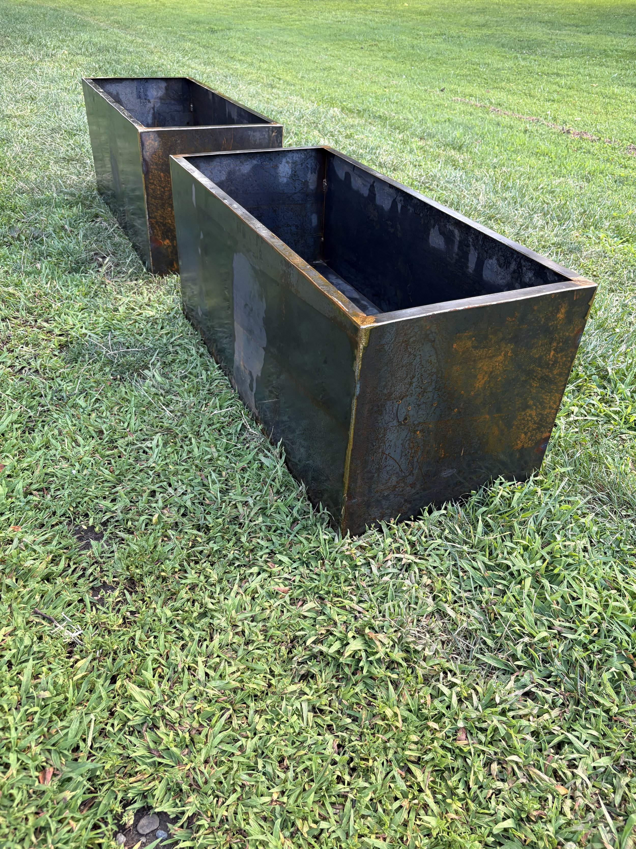 Two large rectangular Corten steel planters placed on a grassy field, with the background showing a patchy lawn.