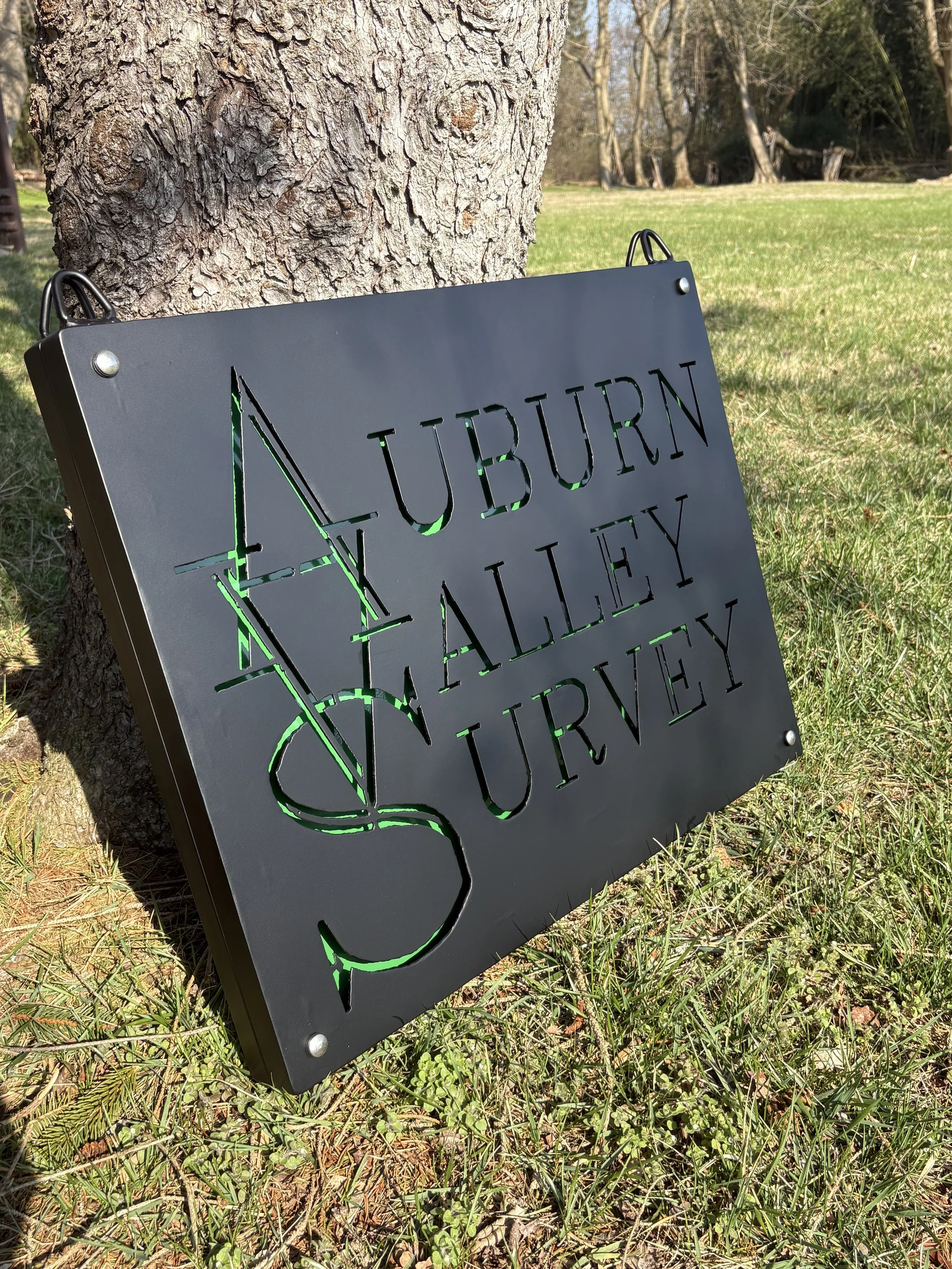A black metal sign with cut-out text that says "AUBURN VALLEY SURVEY" propped against a tree in a grassy park area in Yorklyn DE