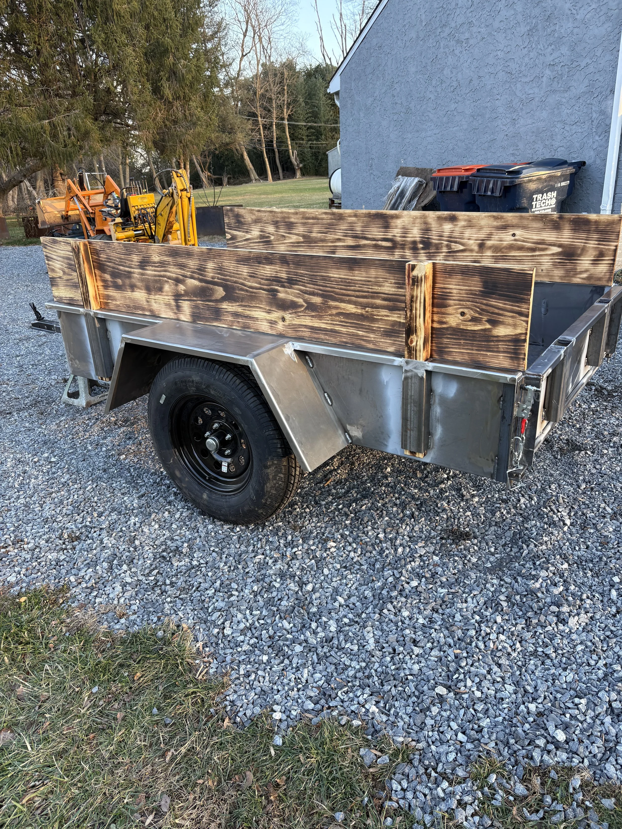 A small trailer with wooden sides, parked on gravel near a gray building, contains outdoor equipment. Trees and a grassy yard are in the background during daylight.