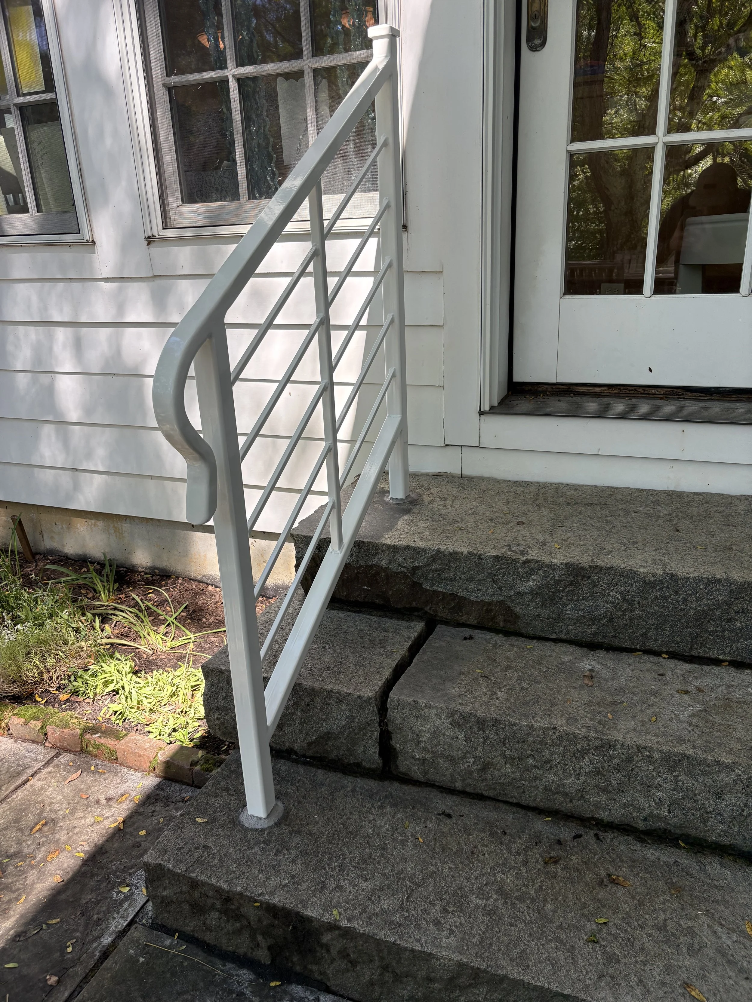 Exterior view of a house entrance showing black-swept concrete steps with a crack, a white railing, a glass-paned door, and part of a window, with plants and trees in the background.