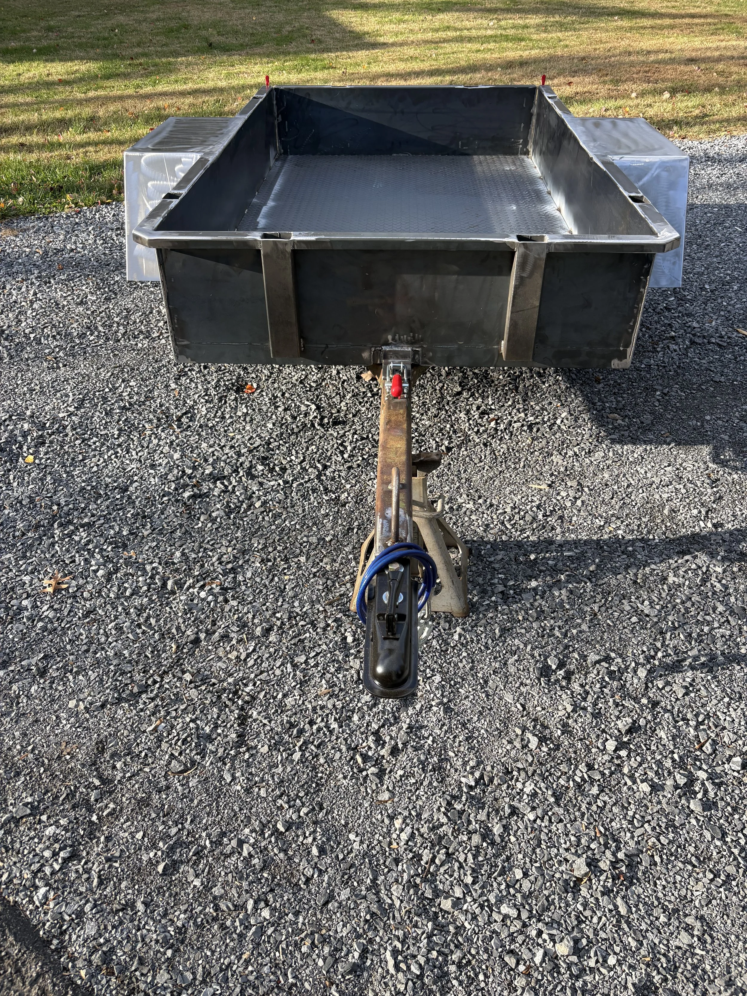 A metal utility trailer with a diamond plate bottom, parked on gravel with grass and trees in the background.