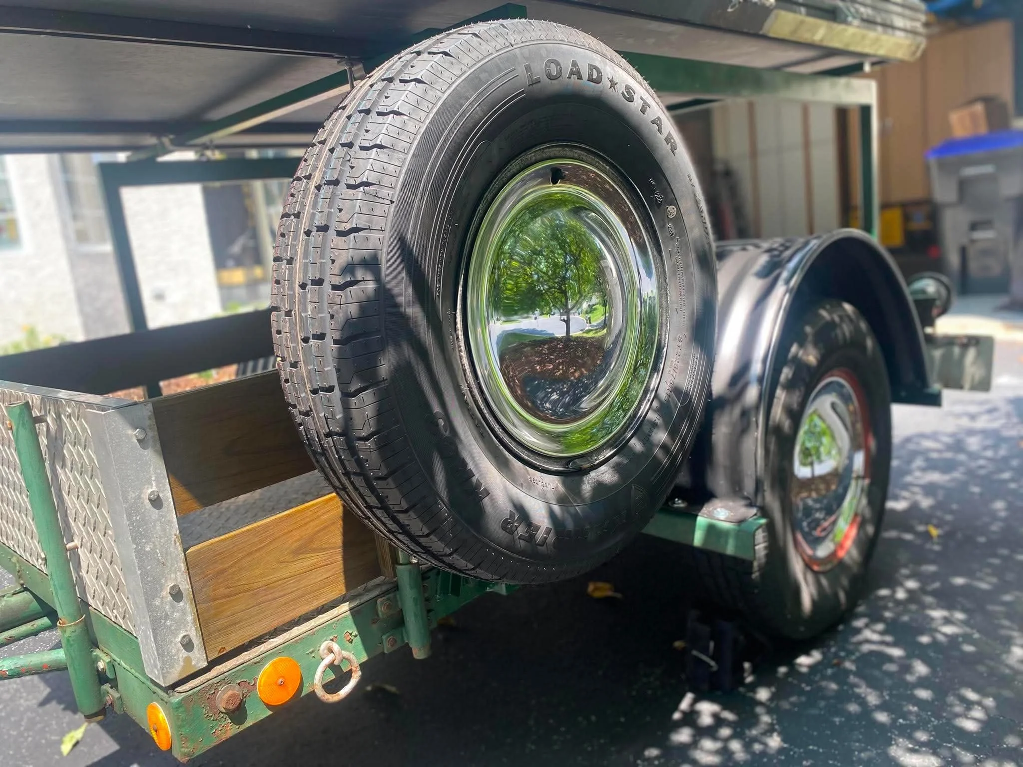 A black tire with the text 'LOAD STAR' on it, mounted on a wheel with a reflective hubcap. The tire is on a flatbed trailer with a wooden deck and green metal structure, parked outside with greenery reflected in the hubcap.