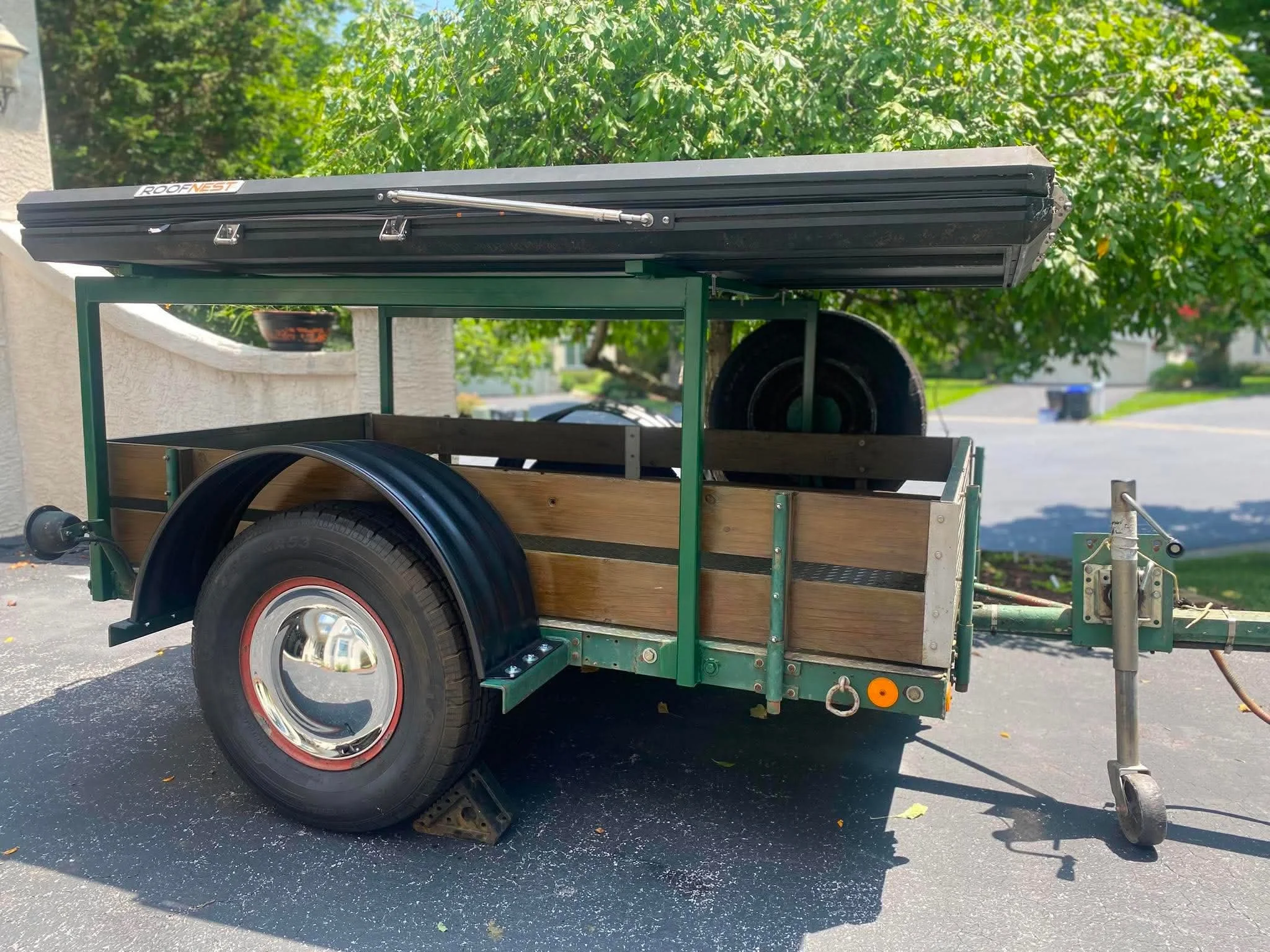 A green utility trailer with a black rooftop tent parked on a residential street. The cart has a tire and a metal frame, with a wooden side panel. In the background, there are trees and a house in Drexel Hill PA