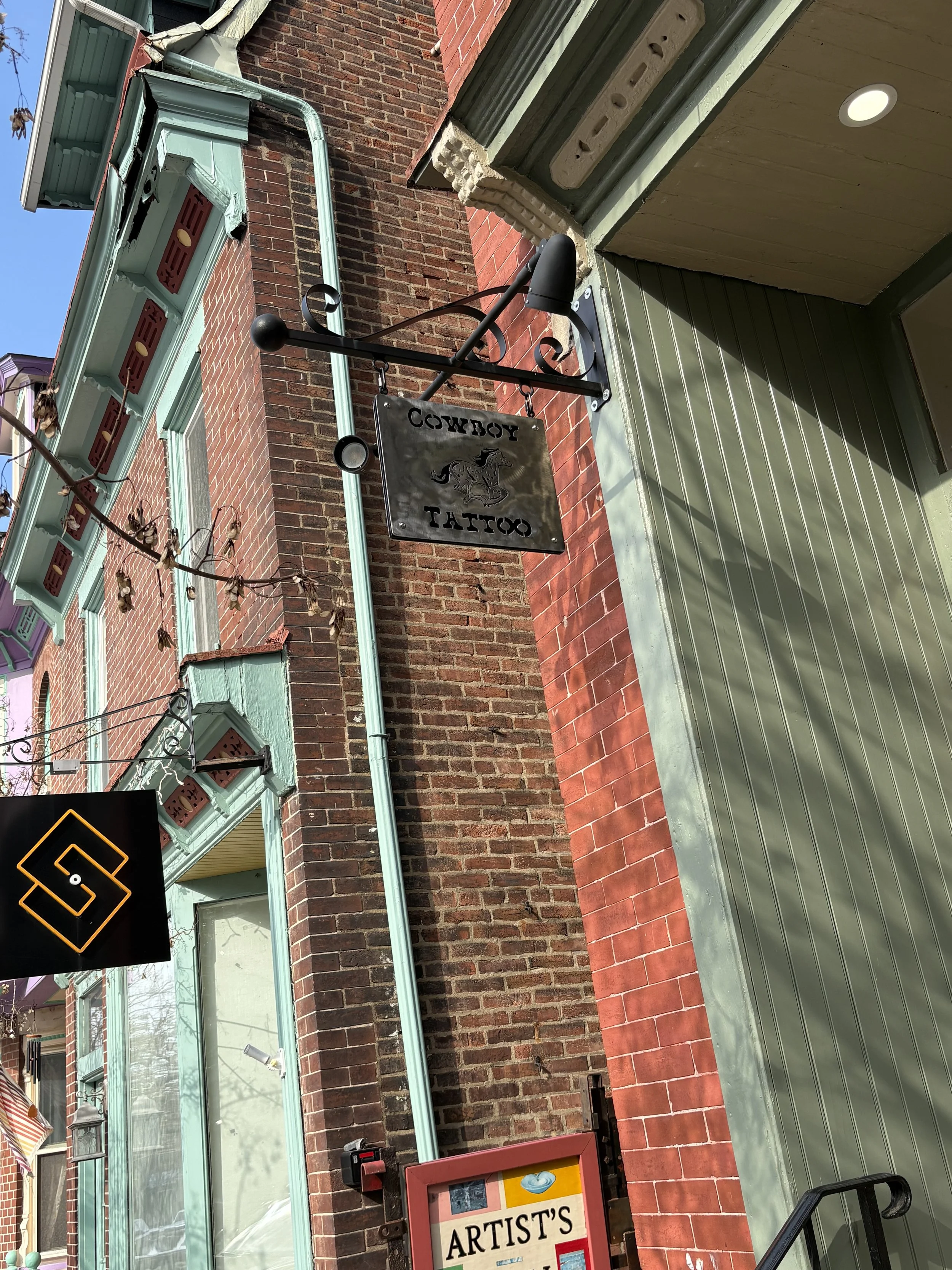Brick building with turquoise window frames and a green roof, a hanging metal sign with a horse, the sign says 'COWBOY TATTOO' hanging in Phoenixville PA