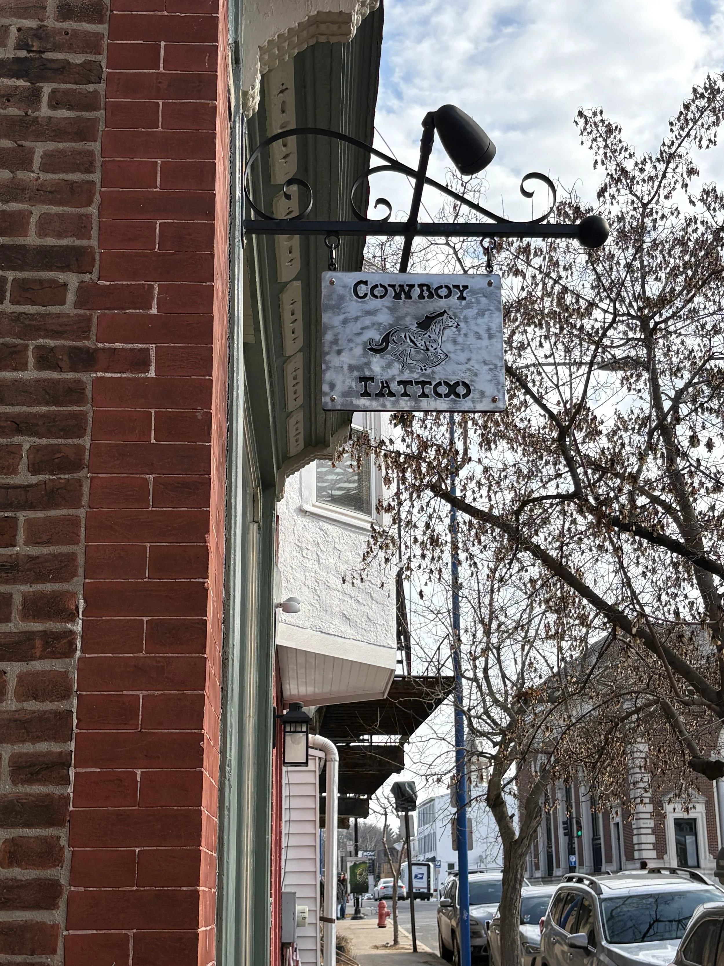 A hanging sign that reads 'Cowboy Tattoo' with a horse graphic, attached to the side of a red brick building, with trees and parked cars on the street in the background.