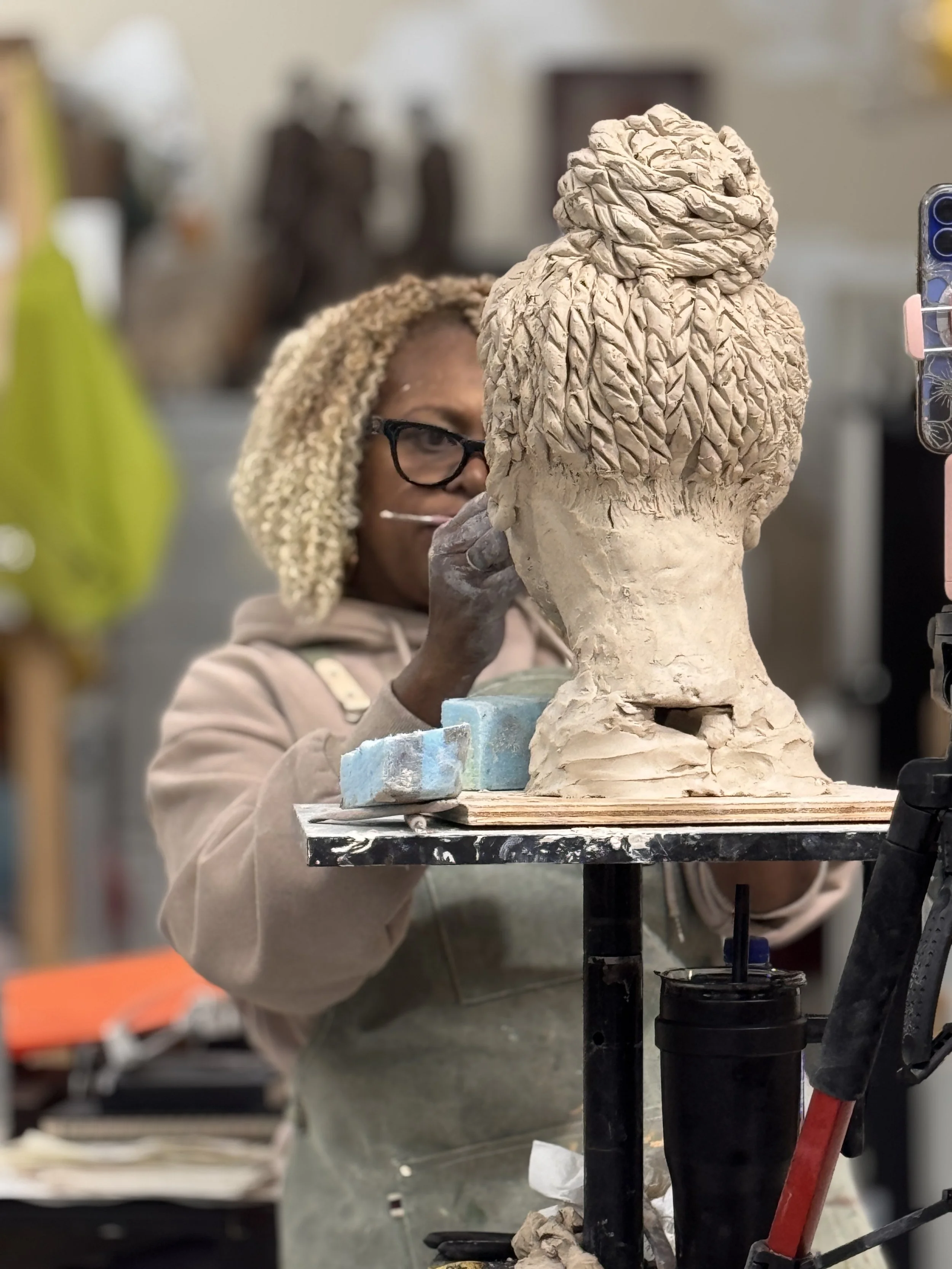 A person working on a sculpture in an art studio, with a partially finished clay bust of a person with textured hair.