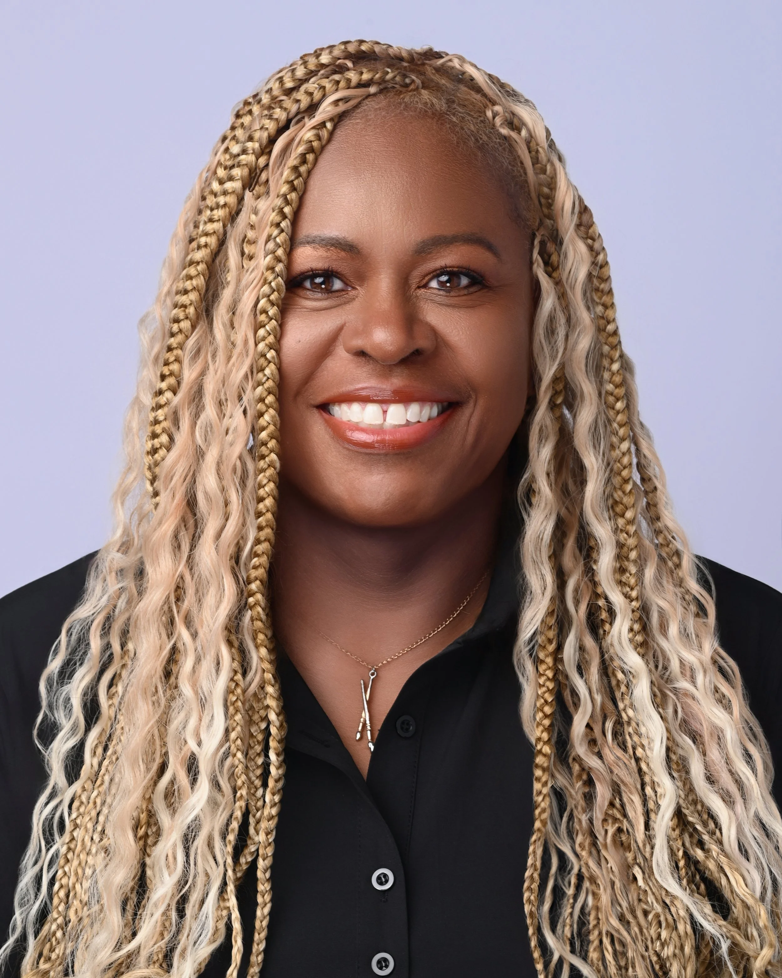 Close-up of a smiling African American woman with long blonde and light brown braided hair, wearing a black shirt and a delicate silver necklace, against a light purple background.