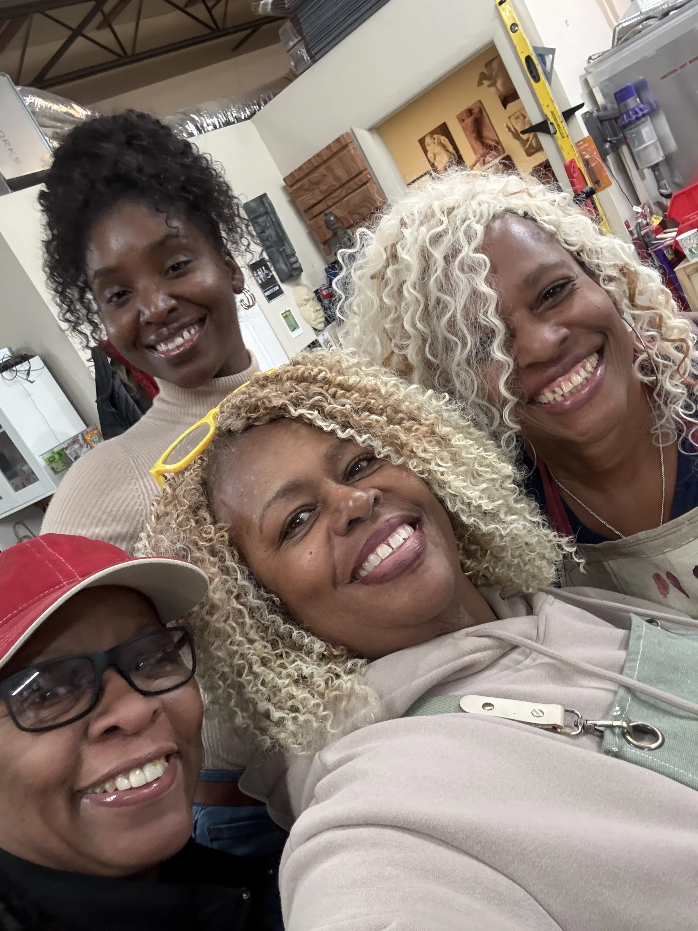 Group of smiling women taking a selfie in a room with kitchen cabinets and tools in the background.