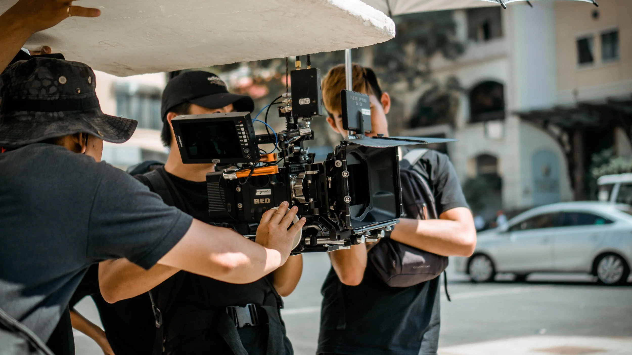 Filmmakers operating a professional camera on an outdoor set with city buildings and parked cars in the background.