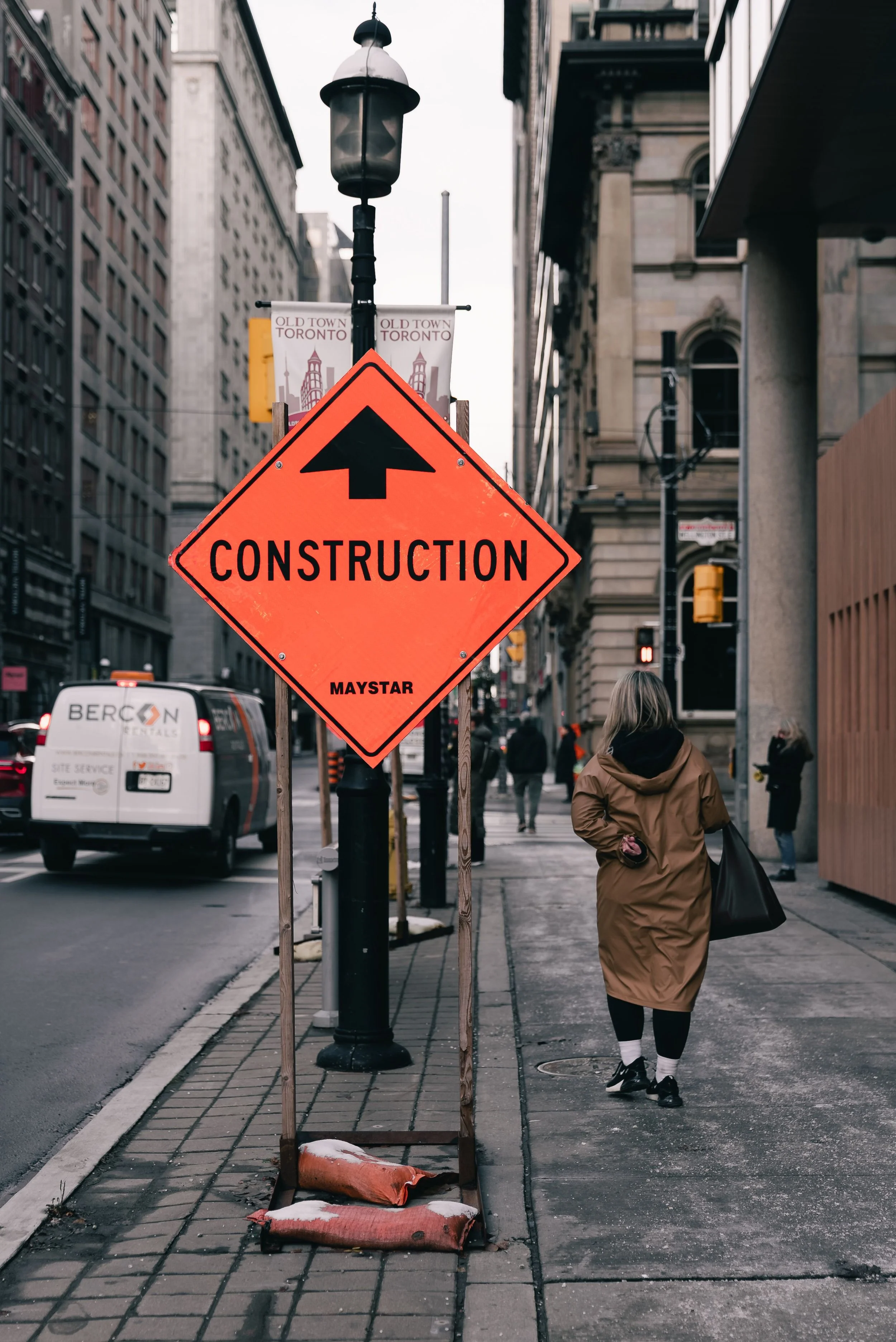 Street view in an urban area showing a bright orange construction warning sign with an upward arrow and the word 'CONSTRUCTION', placed on a sidewalk with sandbags, with pedestrians walking and vehicles in the background.