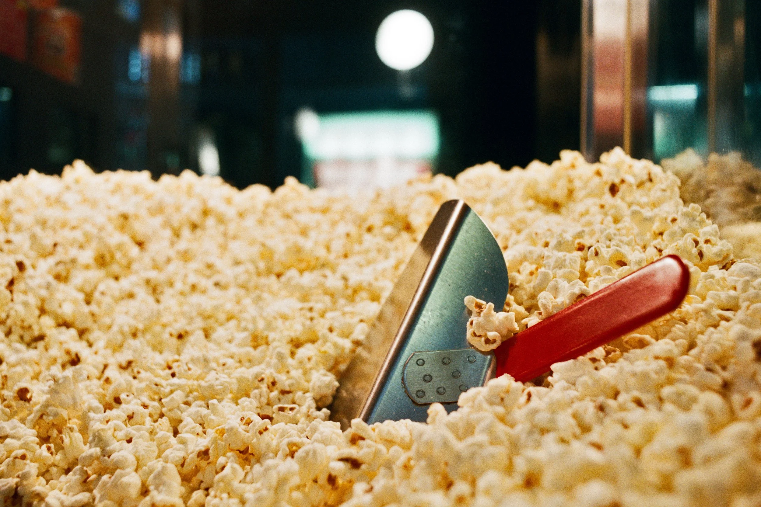 A large container filled with freshly popped popcorn, with a metal scoop resting on the edge, in a movie theater or concession stand setting.