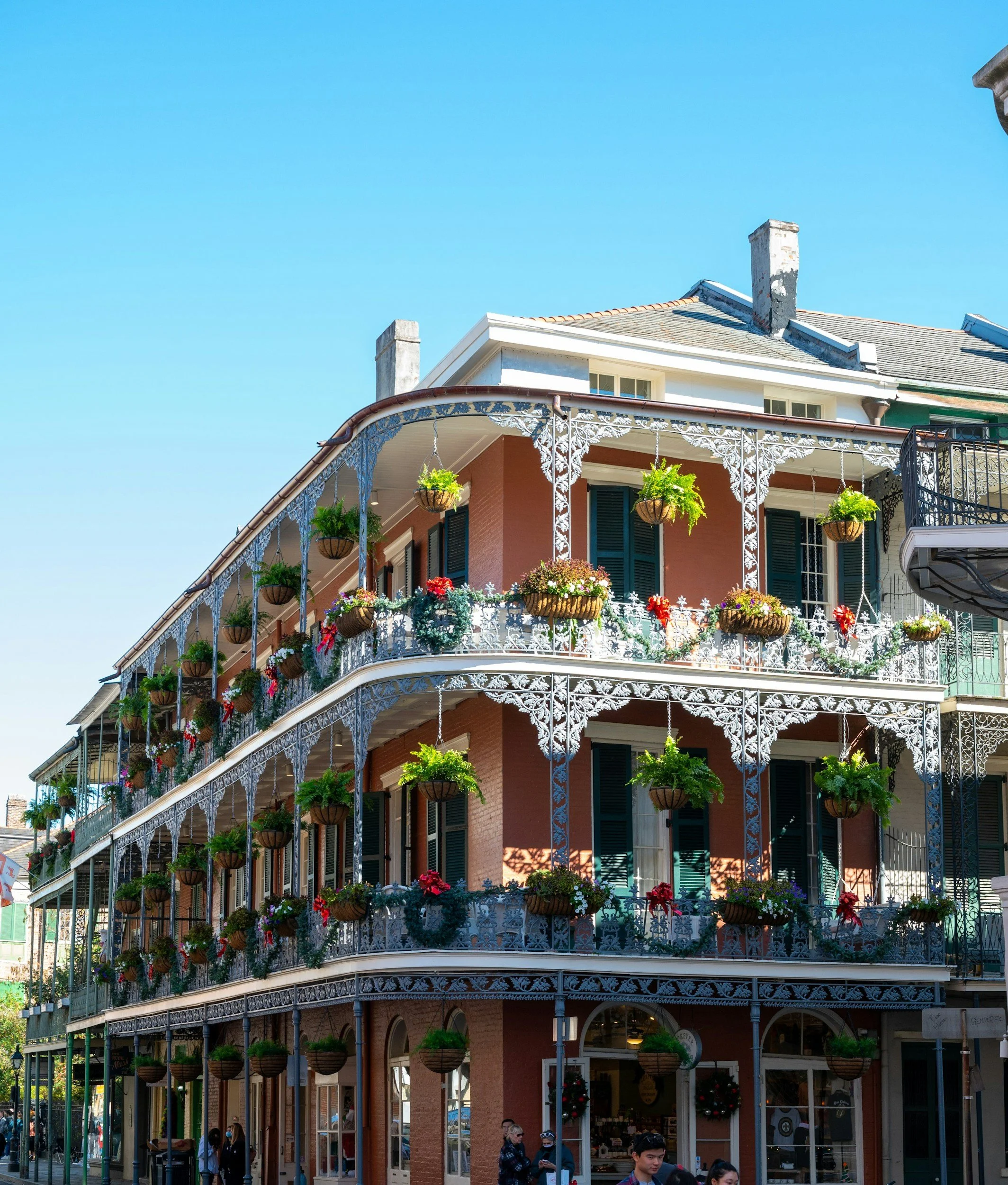 Multi-story building with ornate white iron balcony railings and hanging flower baskets, people walking and shopping outside, bright and sunny day.