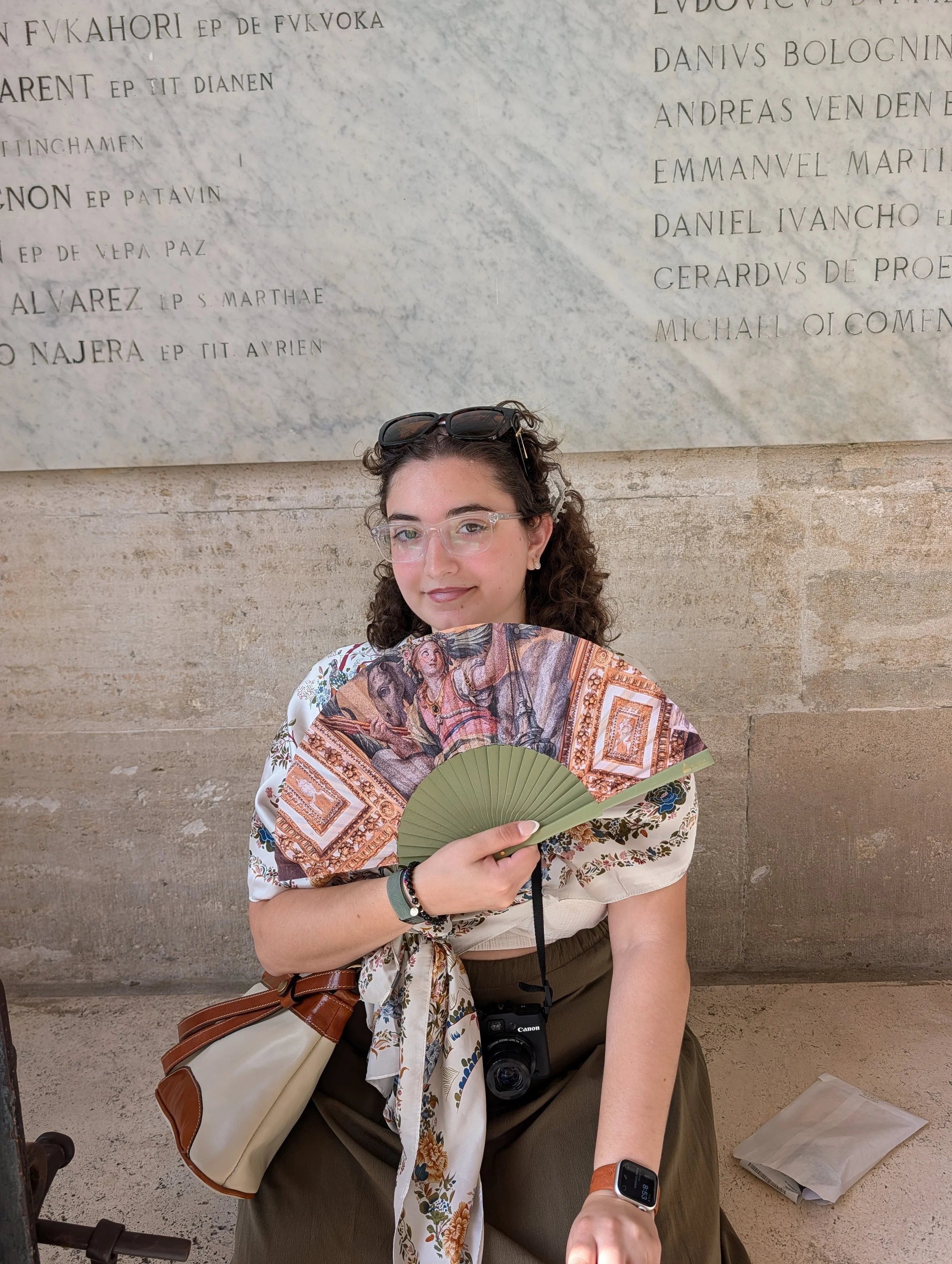 A young woman with curly brown hair, glasses, and a floral scarf holding a decorative fan, seated against a beige stone wall with a large engraved marble plaque behind her. She is wearing an Apple Watch, a bracelet, and a beige and brown handbag on her lap.