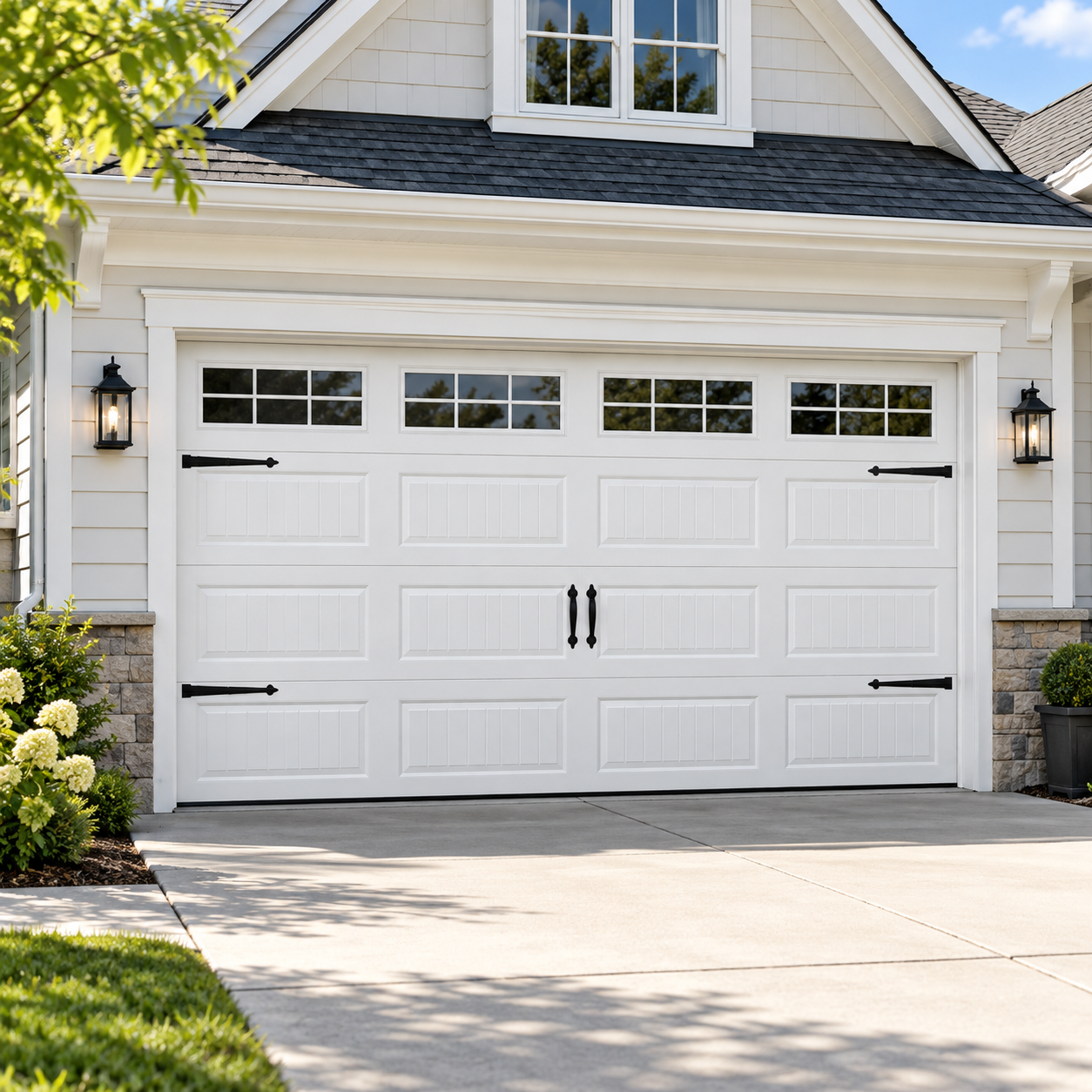 White garage door with black handles and hinges, two wall-mounted lantern lights on either side, and a house with light-colored exterior siding and a gable roof.
