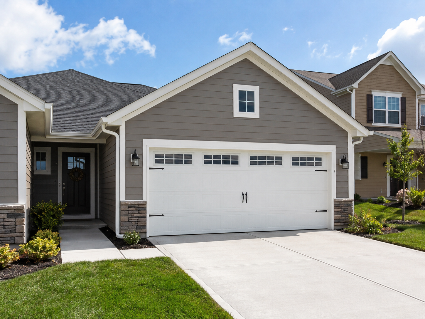 Front view of a suburban house with a gray exterior, white garage door with windows, and small window above the garage. Front yard has green grass and small bushes, with a sidewalk leading to the front door. Clear blue sky with some clouds.