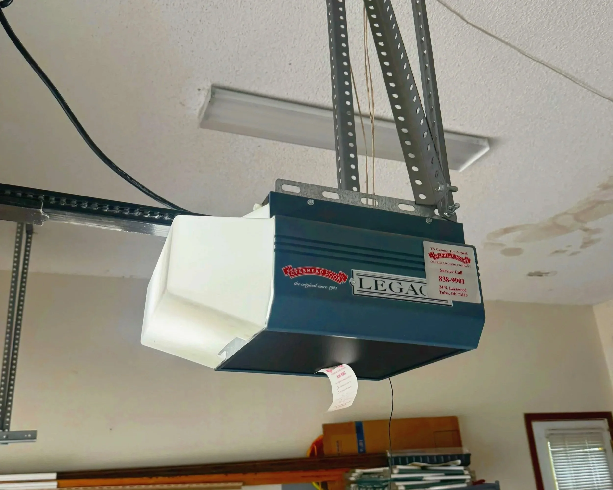 Close-up of a garage door opener motor with a metal rail and an emergency release cord hanging from it, mounted on a ceiling with exposed beams and a drop ceiling tile.
