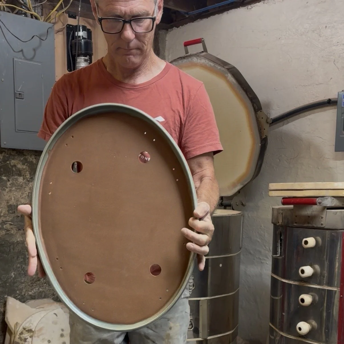 A man wearing glasses and a red t-shirt holding an oval-shaped pottery piece with a brown interior, standing in a workshop with equipment including a large circular kiln and pipes.