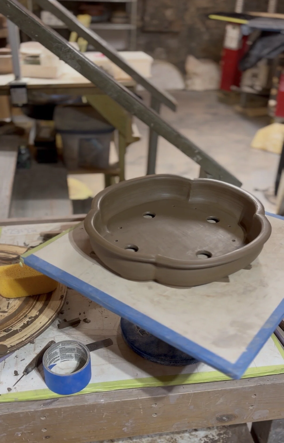 A pottery wheel with a partially formed mokka bonsai pot set on a drywall board in a pottery studio. Various tools, tape, and equipment are visible in the background.