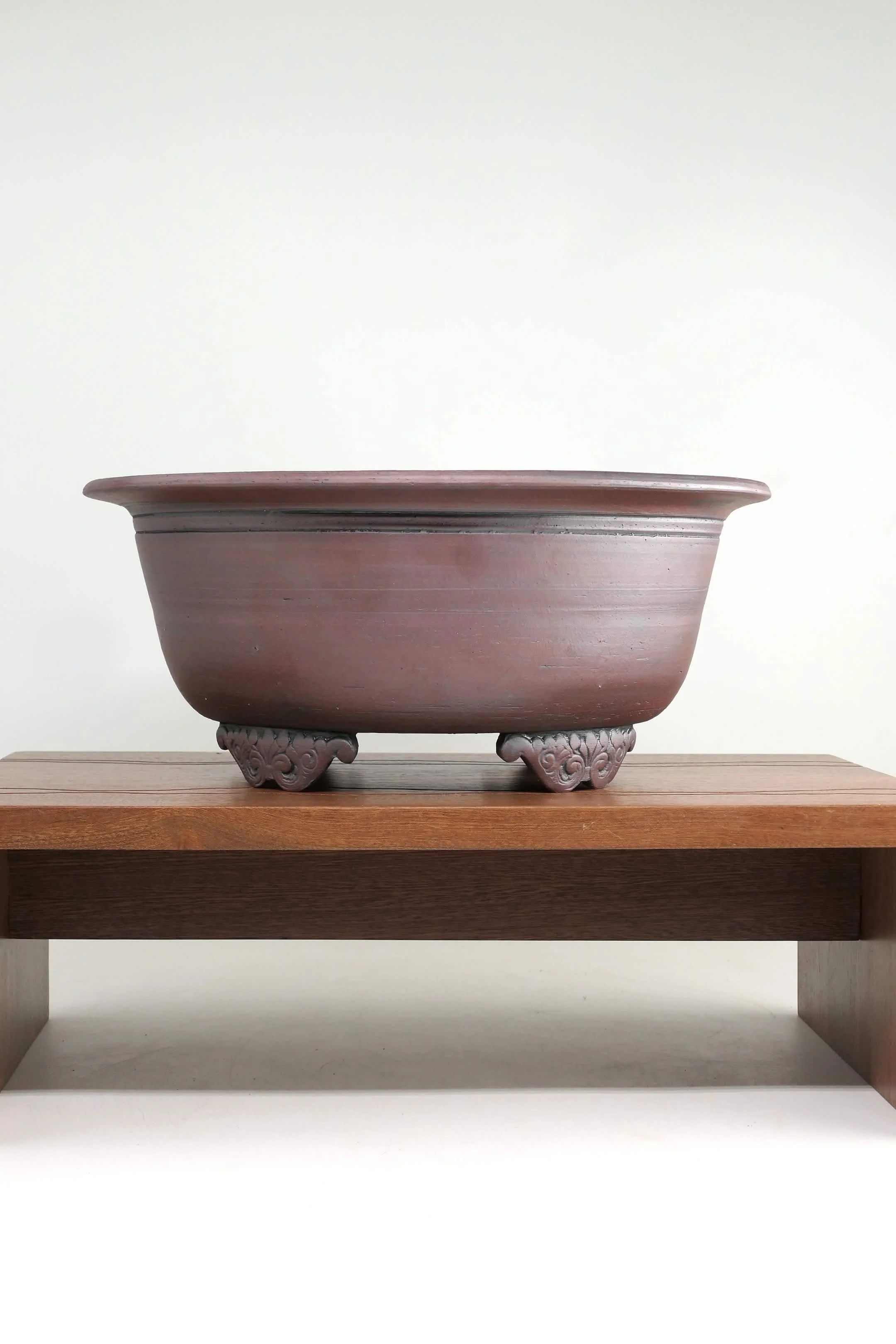 A large, round, reddish-brown ceramic bonsai pot with a wide rim, resting on ornately decorated feet, placed on a wooden surface against a plain white background.