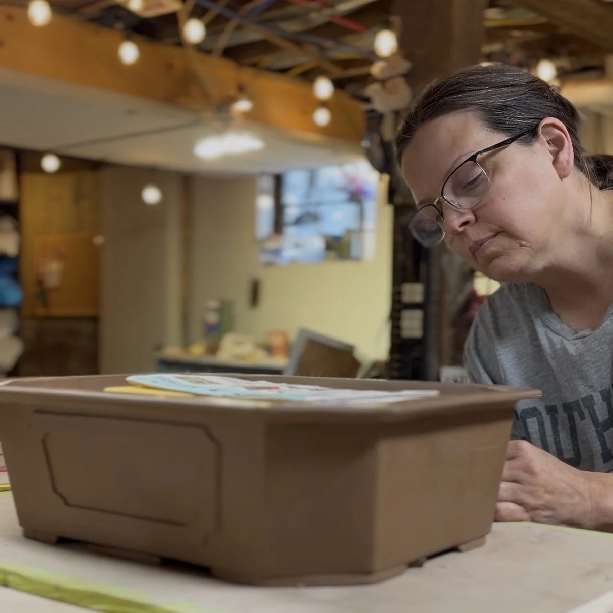 Sharon Fox wearing glasses leaning over a rectangular bonsai pot in a workshop or craft space, with wooden shelves and tools in the background.
